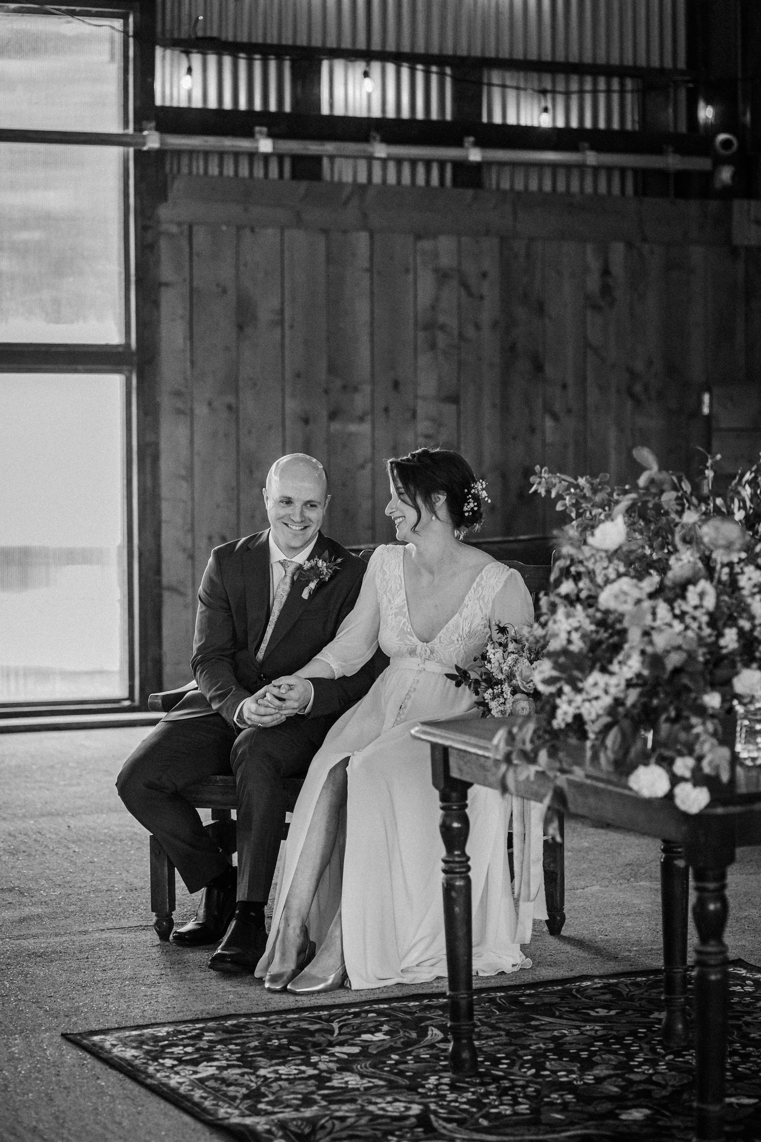 Black and white photo of a smiling bride and groom seated together during their rustic barn wedding ceremony.