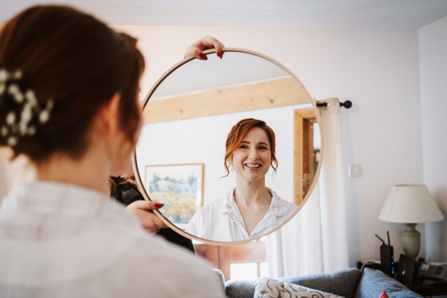 Bride smiling at her reflection in a round mirror while getting ready on her wedding day.