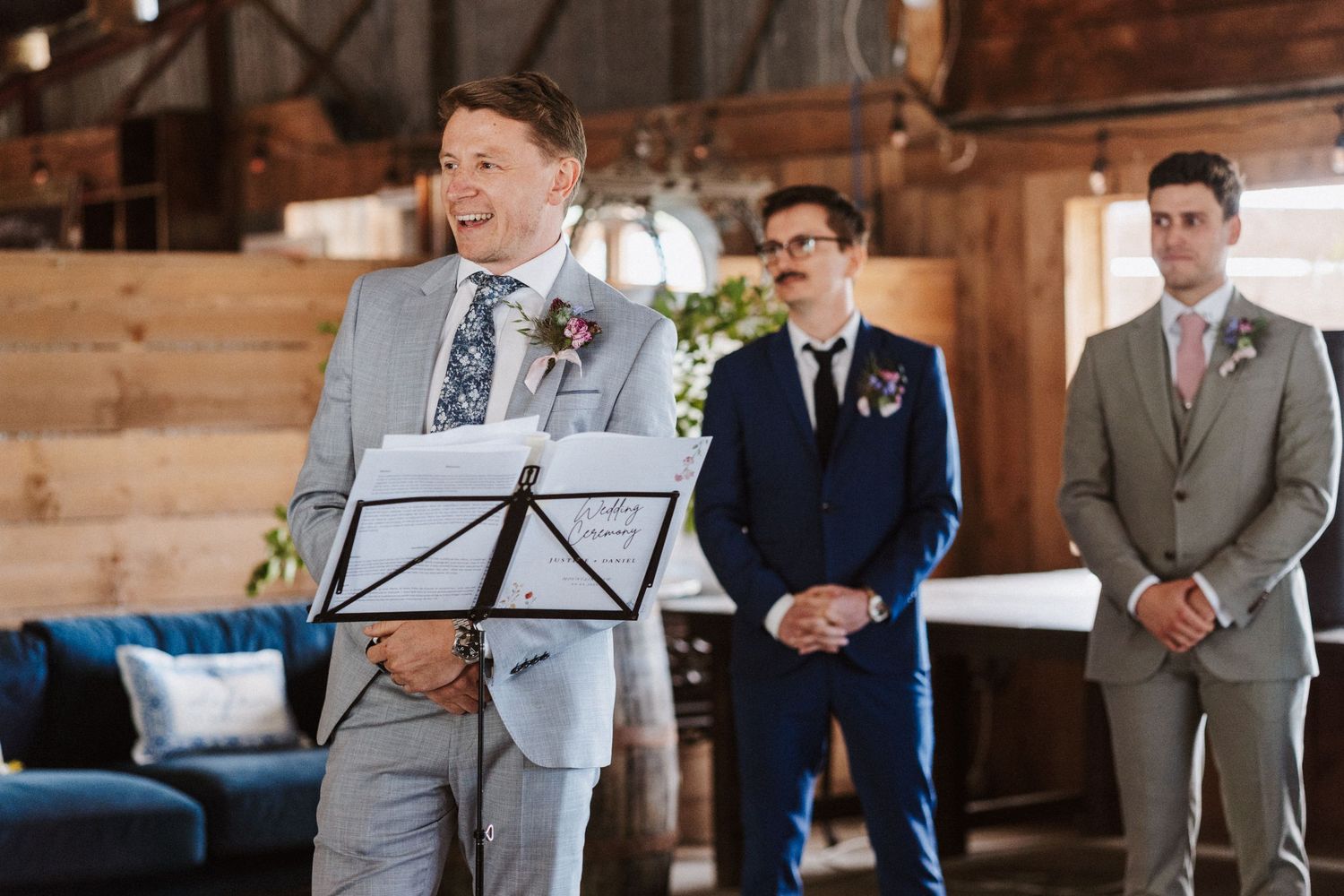 Man in light gray suit speaking at wedding podium with groomsmen standing behind him in rustic barn venue.