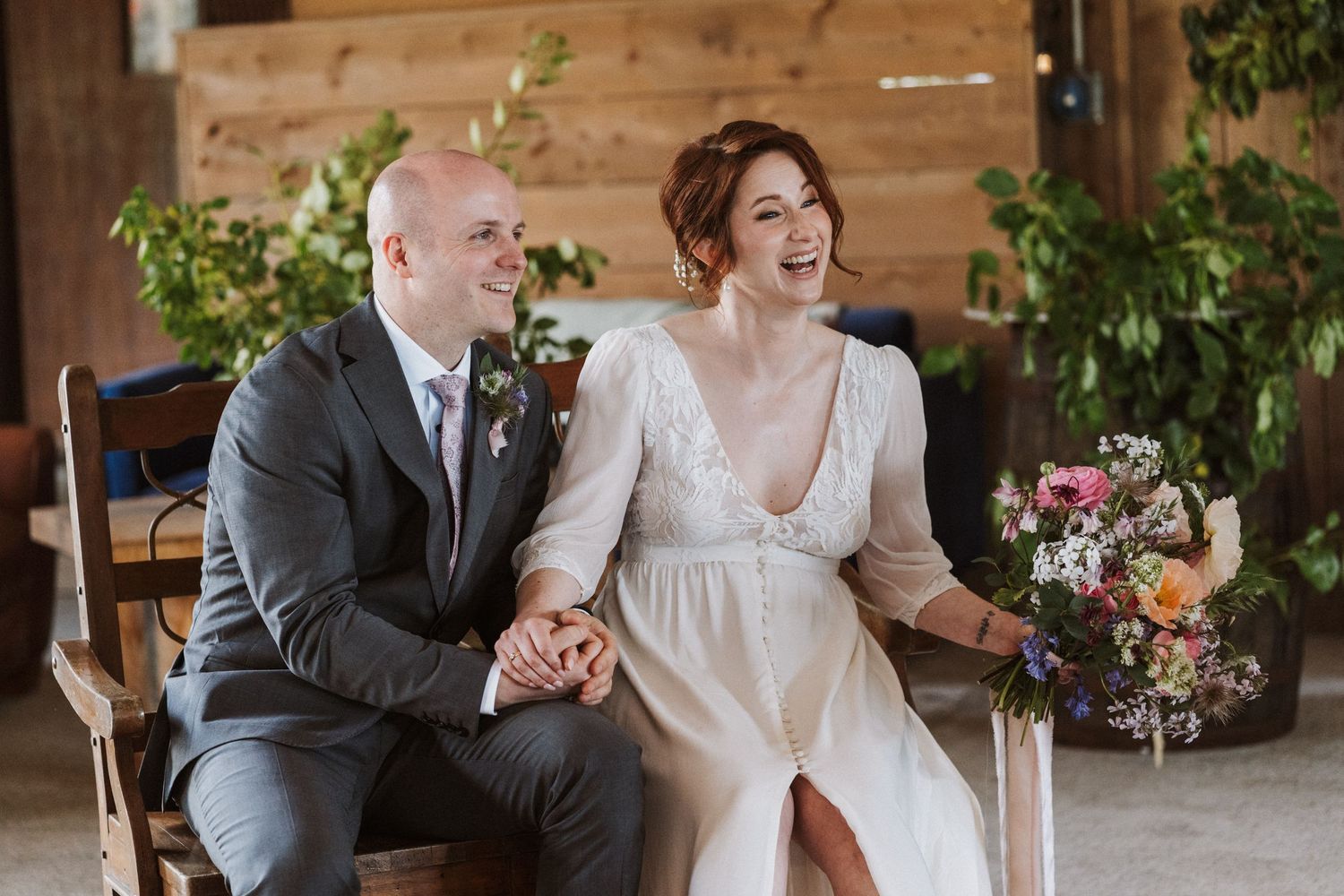 A laughing bride in a white gown and groom in gray suit sit together holding hands at a rustic barn wedding.