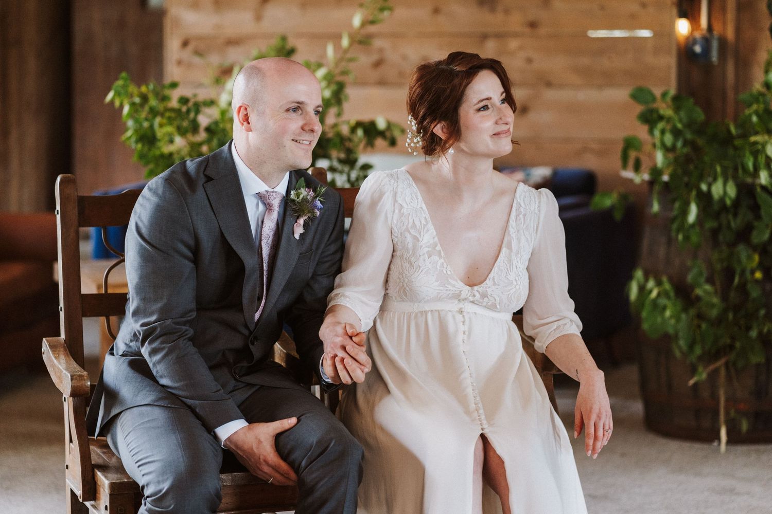 Bride and groom sit together holding hands, smiling in a rustic barn wedding venue with greenery decor.