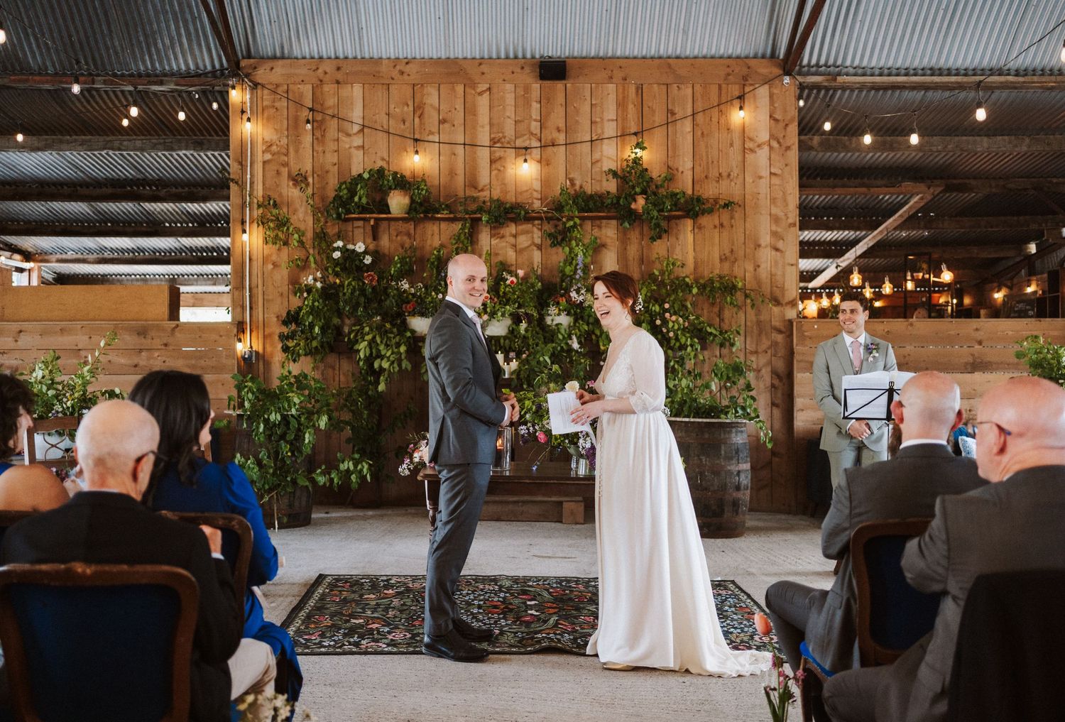 Couple exchanging vows at rustic barn wedding ceremony with wooden wall, greenery backdrop, and string lights.