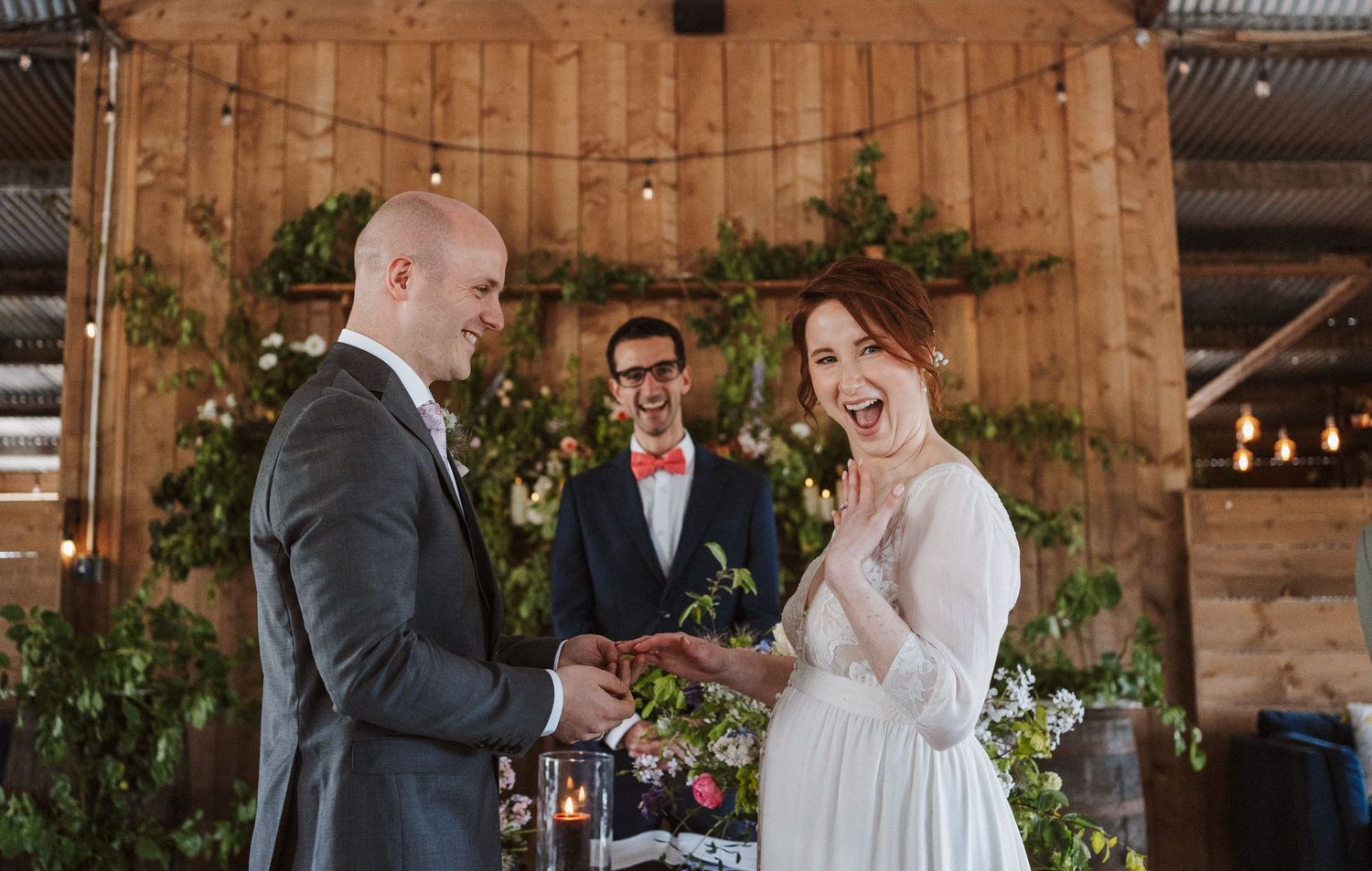 Bride laughs joyfully as groom places ring on her finger during rustic barn wedding ceremony with officiant.
