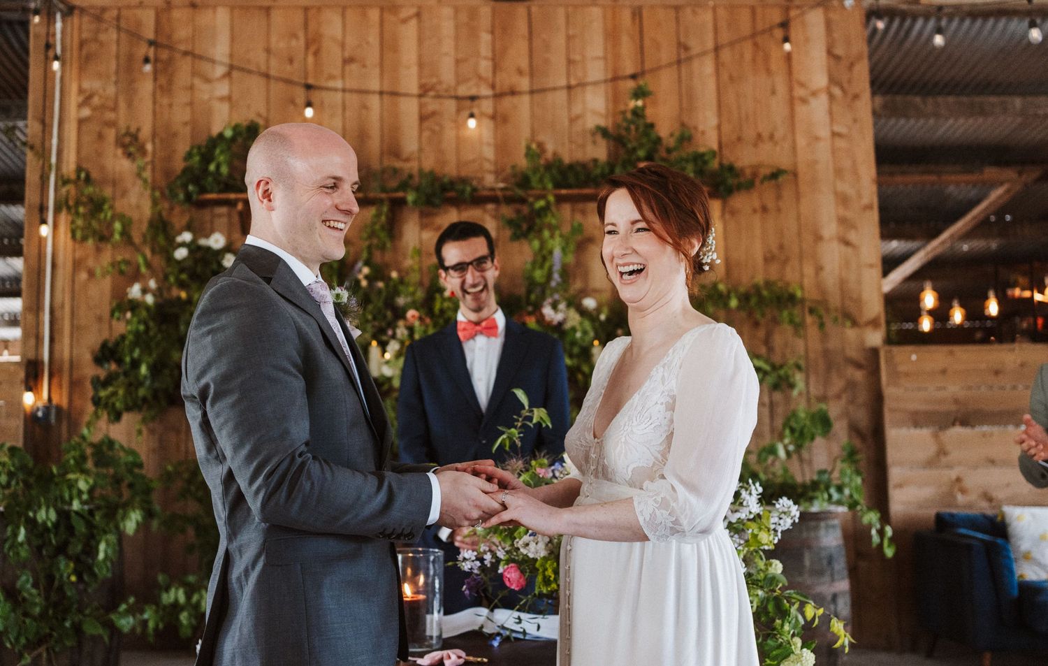 Bride and groom exchanging vows at a rustic barn wedding ceremony with greenery decor and officiant in background.
