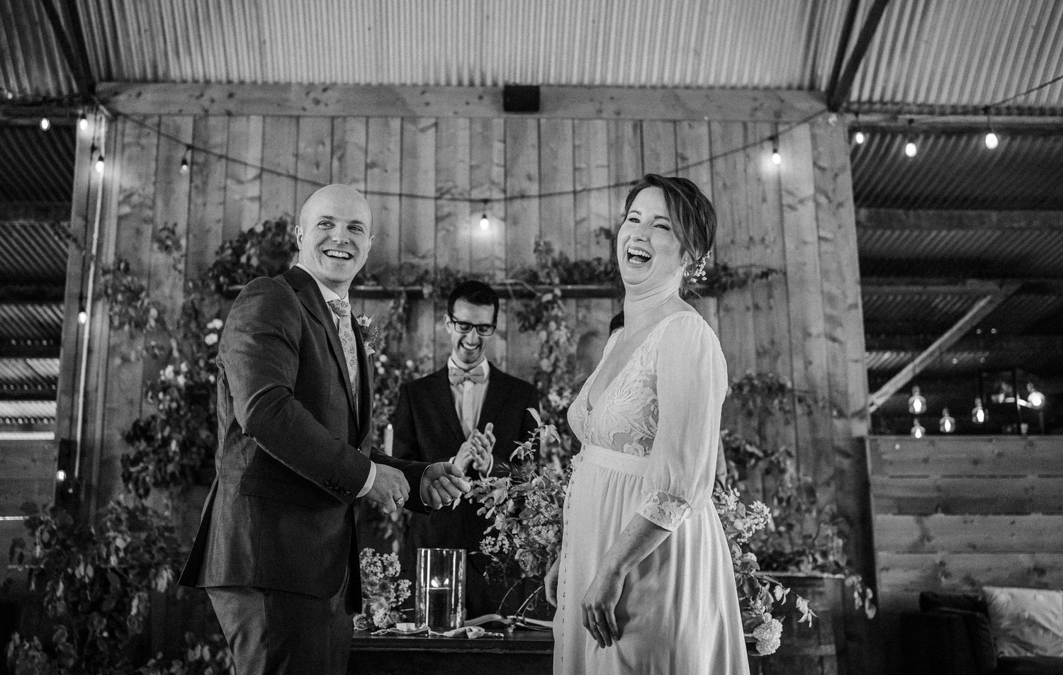 Black and white photo of a laughing bride and groom shaking hands with an officiant at a rustic barn wedding ceremony.
