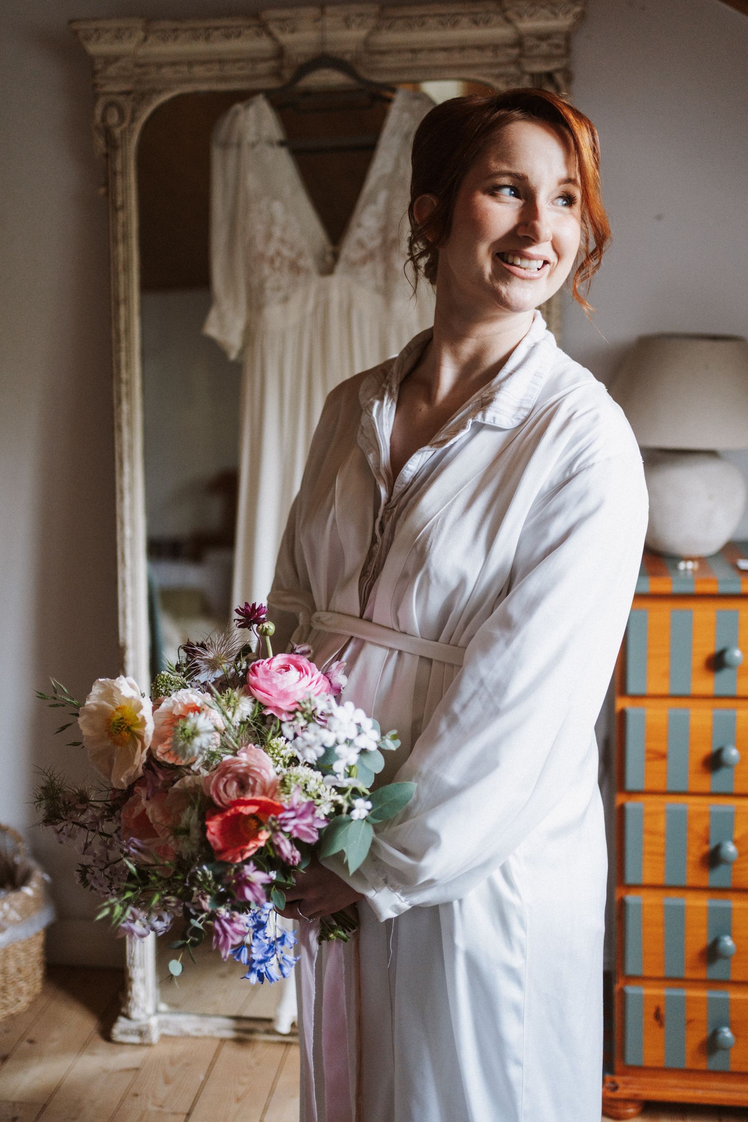 Smiling bride in white robe holding colorful wedding bouquet with pink roses, standing near mirror with gown hanging behind.