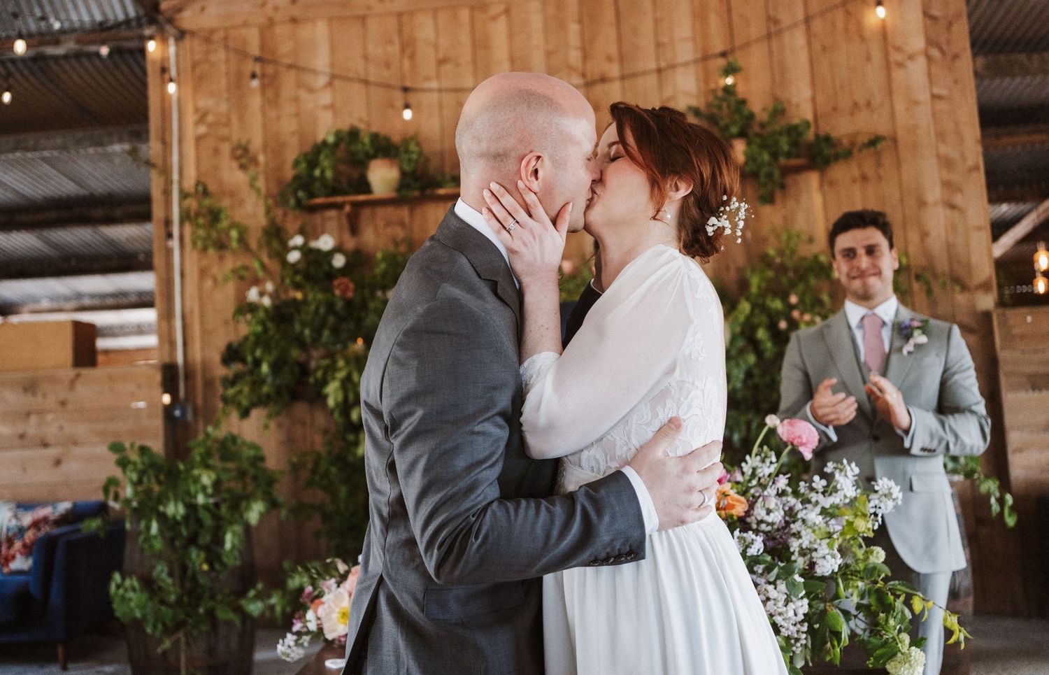 Bride and groom share first kiss at rustic barn wedding ceremony with floral backdrop and string lights.