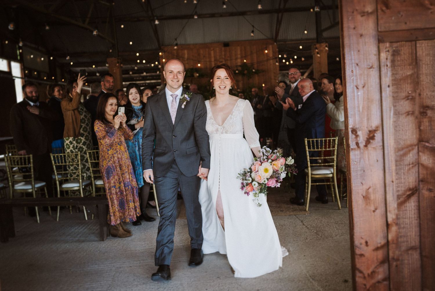 Bride and groom walk down aisle at rustic barn wedding reception as guests applaud and celebrate.