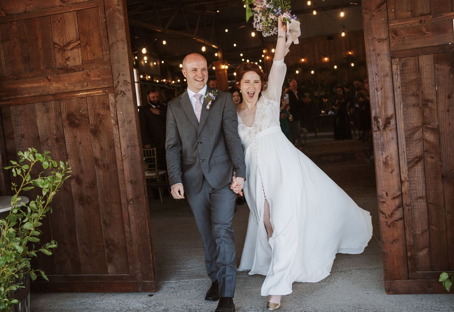 Joyful bride raising bouquet and groom holding hands exiting rustic barn wedding venue with string lights.