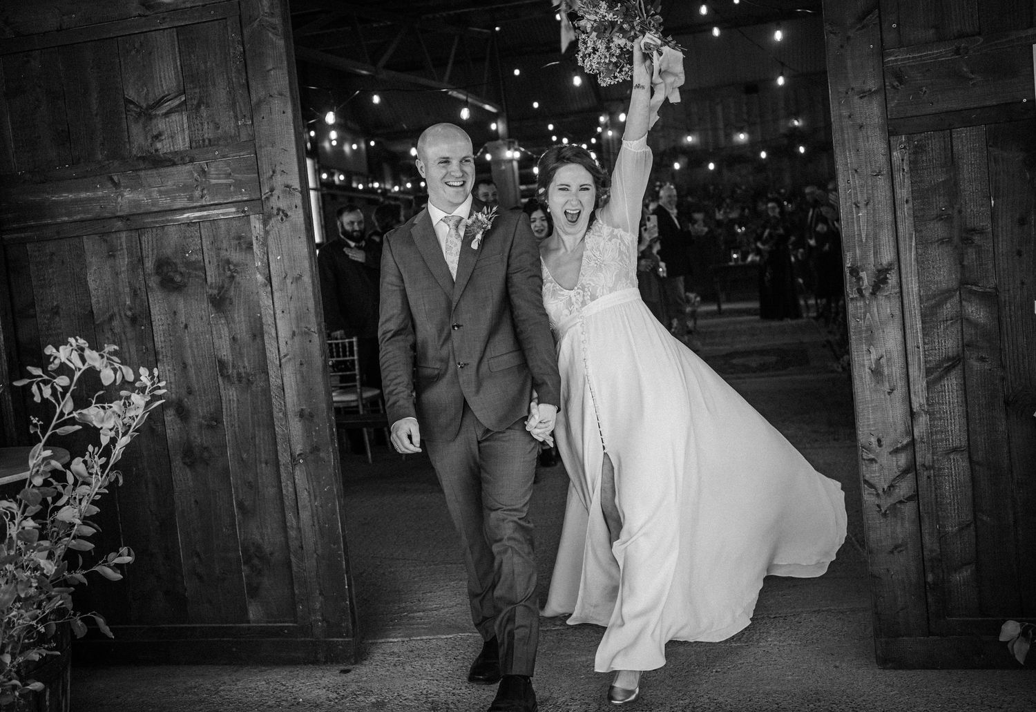 Black and white photo of a joyful bride and groom entering their wedding reception, bride raising bouquet.