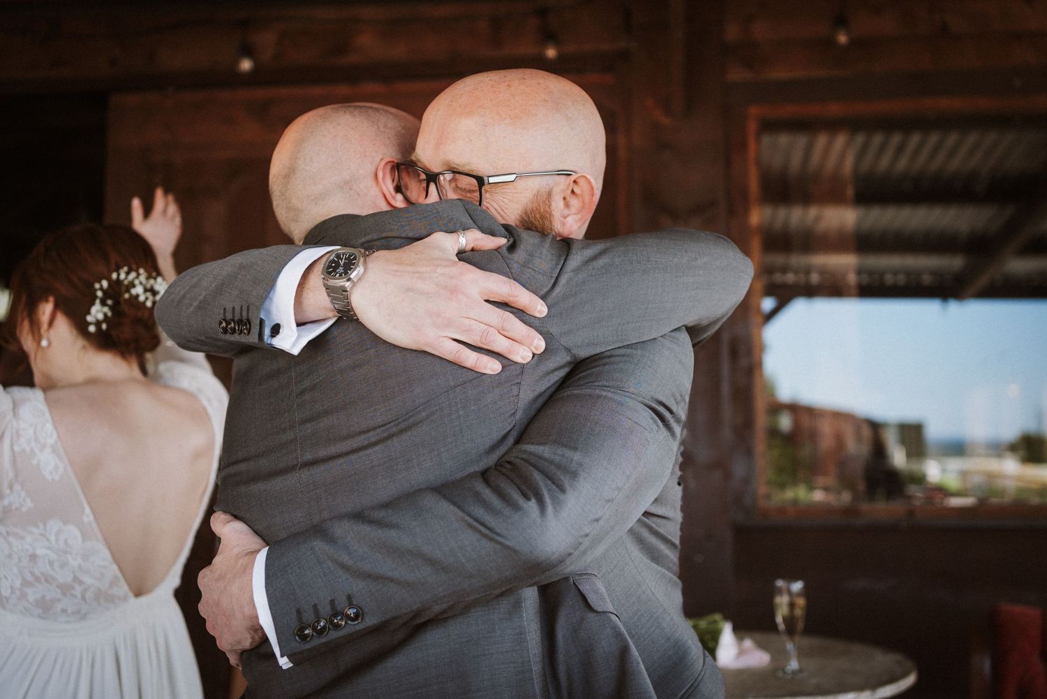Two bald men in suits share an emotional hug at a wedding ceremony, with a bride visible in the background.