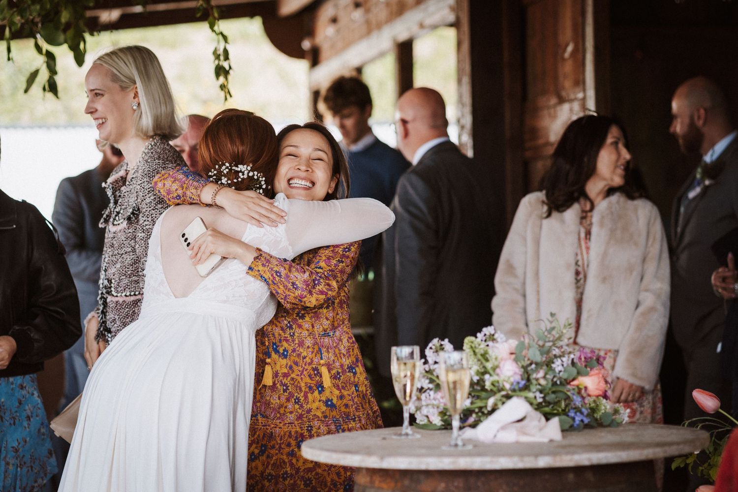 Bride in white dress hugging smiling guest at rustic wedding reception with champagne glasses on wooden table.
