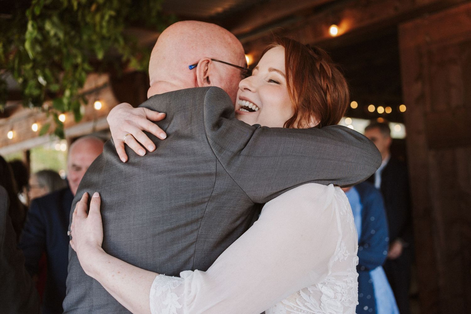 A bride with red hair laughs joyfully while embracing a bald man in a gray suit at a warmly lit wedding reception.