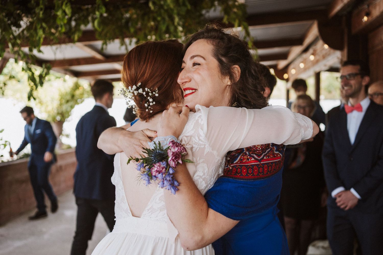 Two women sharing a warm hug at a wedding celebration, one wearing a white bridal gown with floral hair accessories.