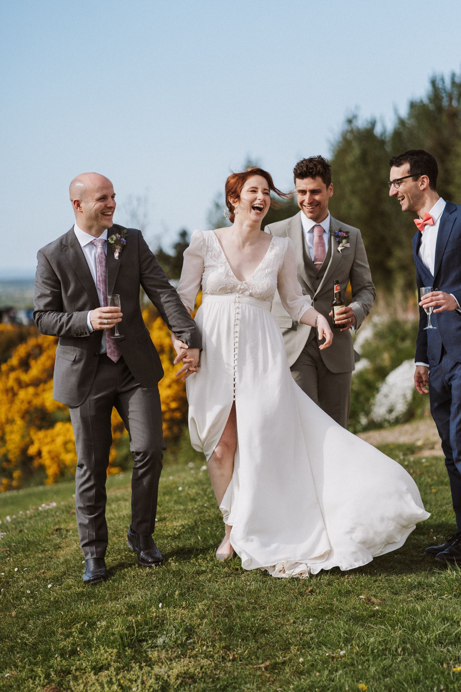 Laughing bride in white gown walks with groomsmen holding champagne glasses outdoors on a sunny wedding day.