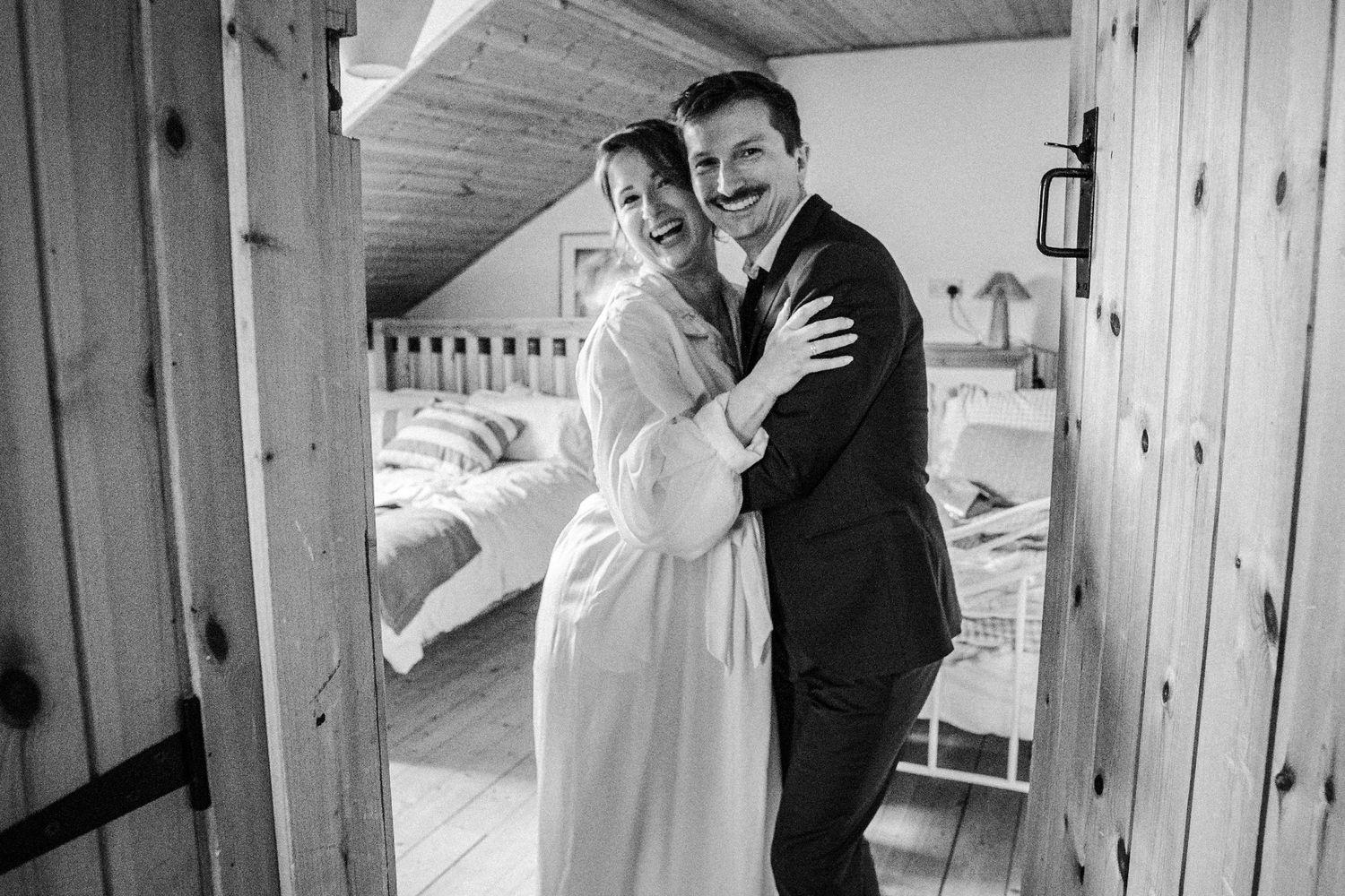 Black and white photo of a smiling couple embracing in a rustic wooden doorway, dressed in wedding attire.