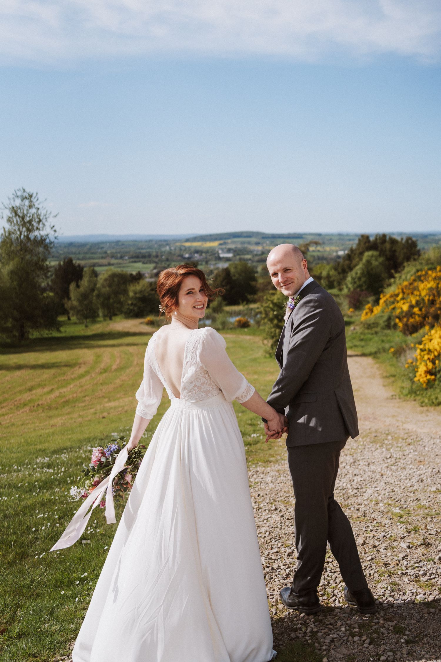 Bride and groom holding hands walking along a gravel path with lush green countryside views on a sunny wedding day.