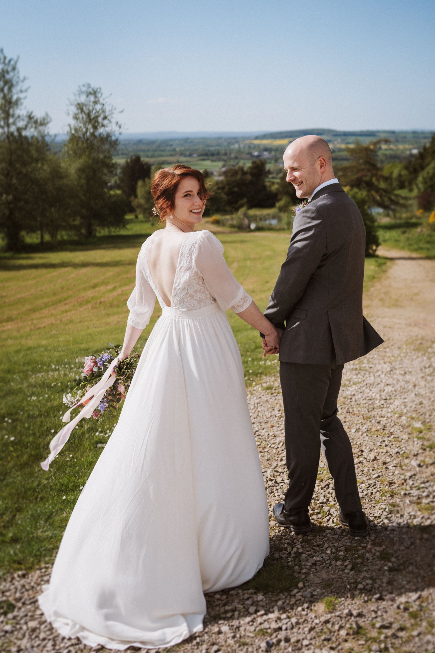 Bride in white lace-back gown holds groom's hand on gravel path, smiling over shoulder with scenic countryside views.