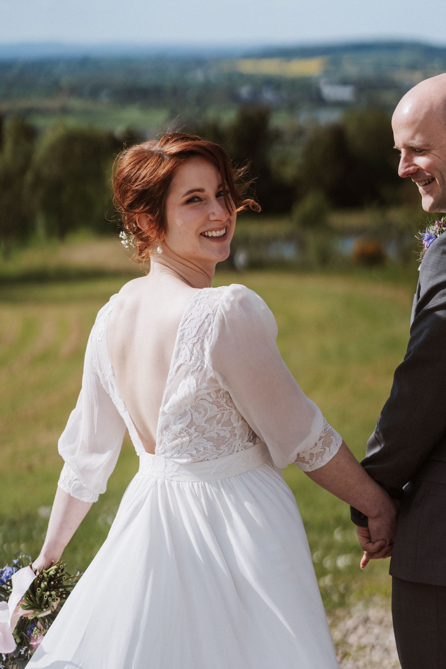 Smiling bride in open-back lace wedding dress holds bouquet while holding groom's hand outdoors with green landscape.