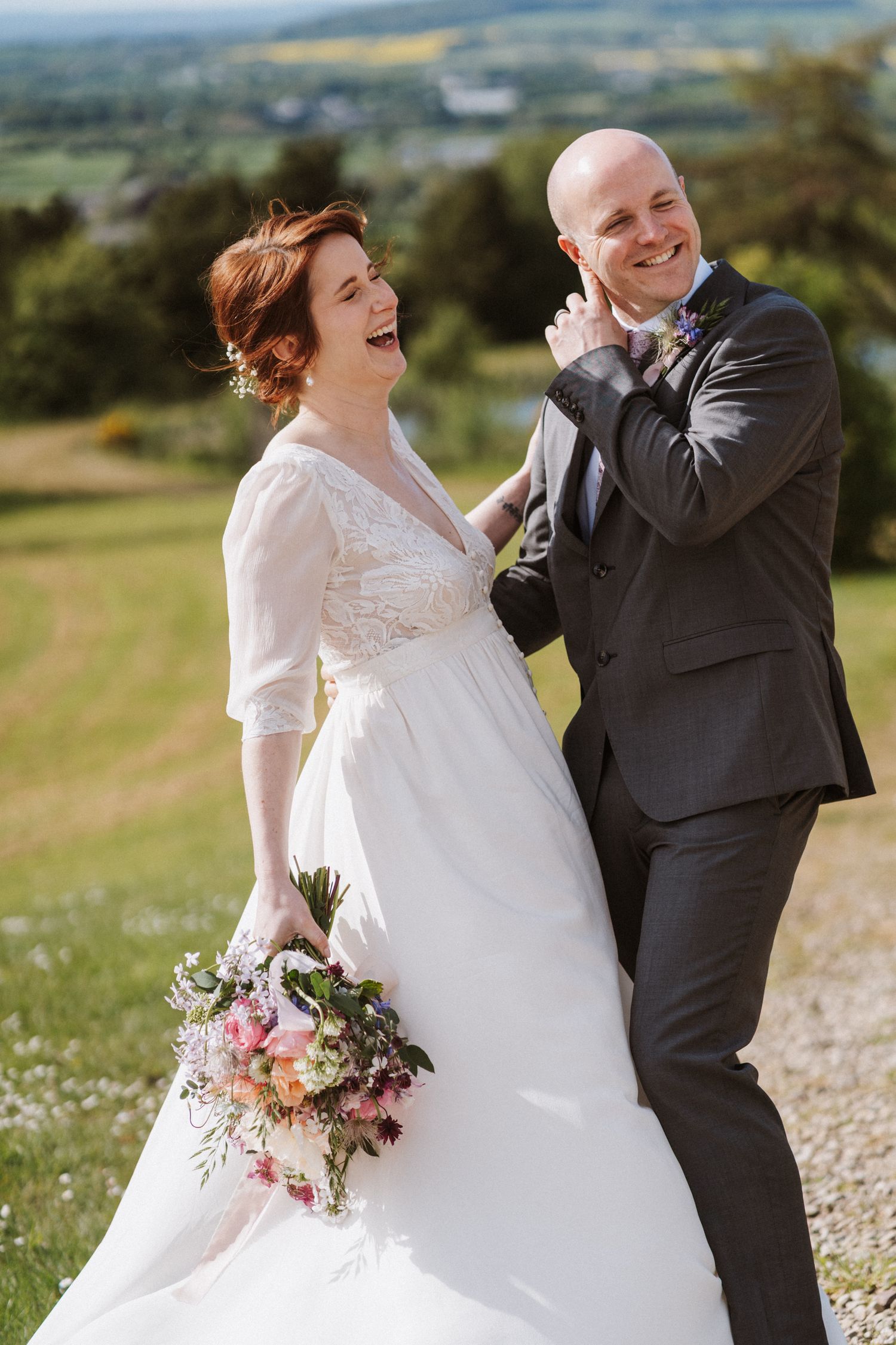 Laughing bride in white lace gown holding bouquet with smiling groom in dark suit outdoors on sunny wedding day.