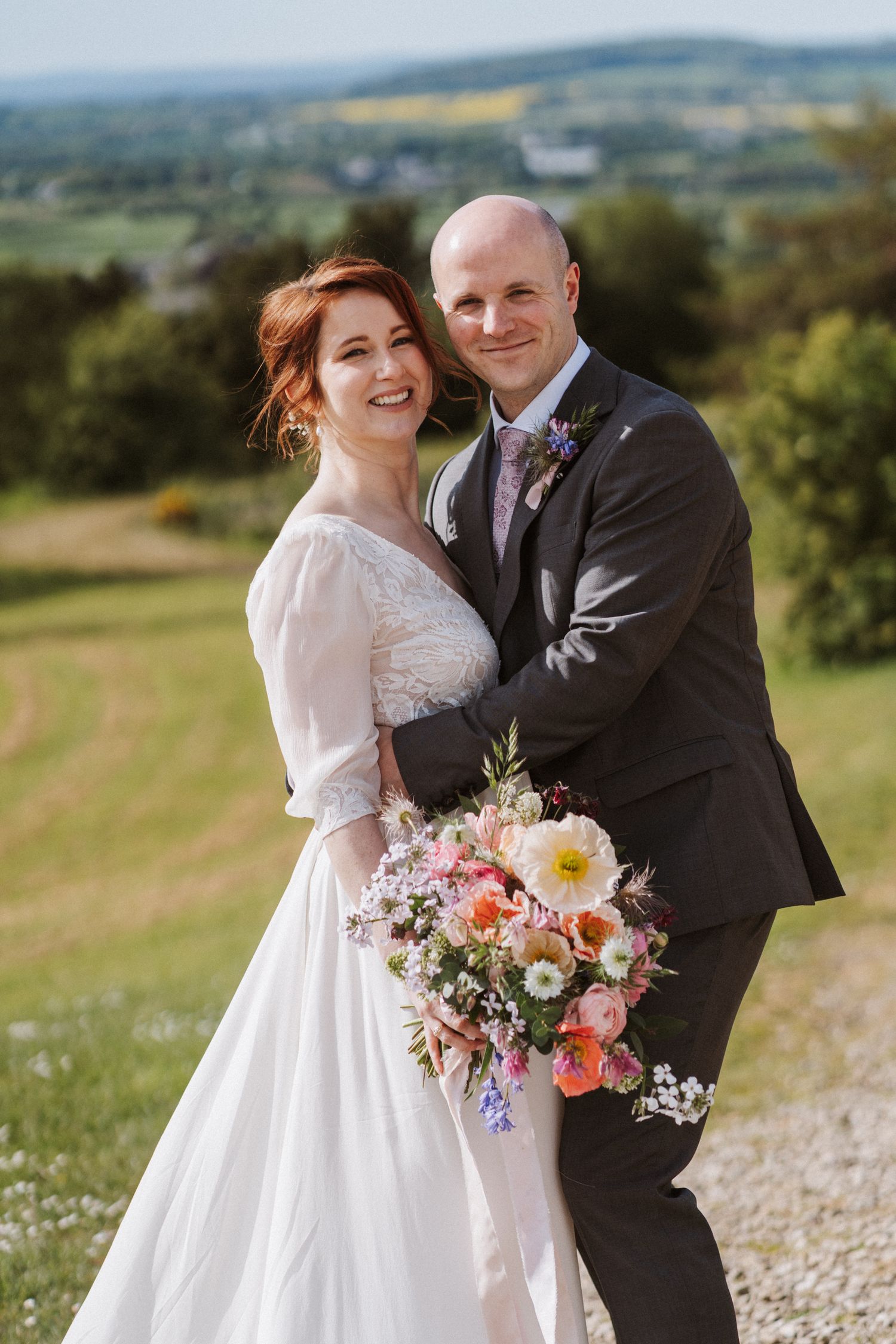 Bride and groom pose outdoors holding a colorful bouquet with rolling countryside views behind them on their wedding day.