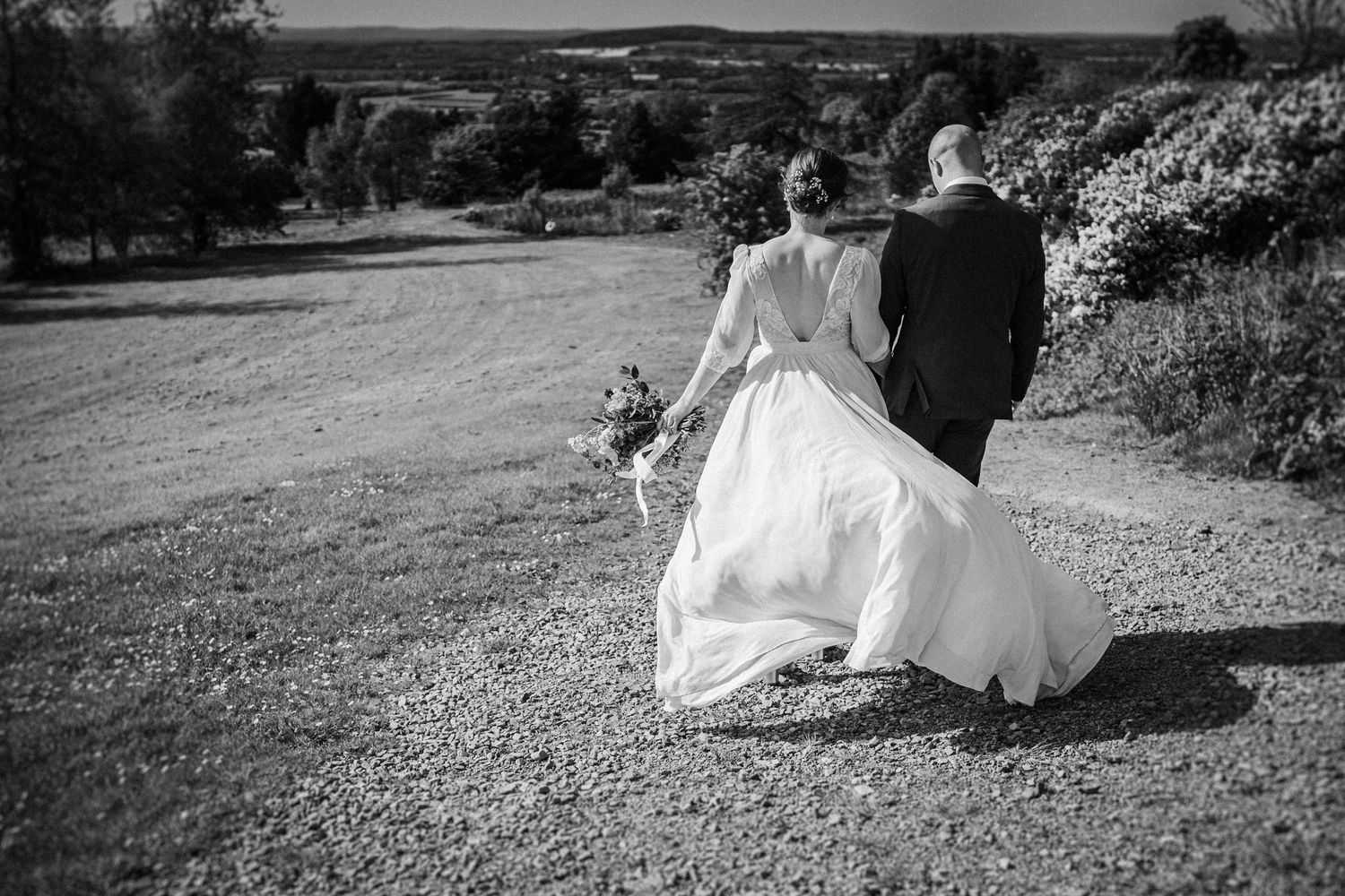 Black and white photo of a bride and groom walking hand in hand along a gravel path on their wedding day.