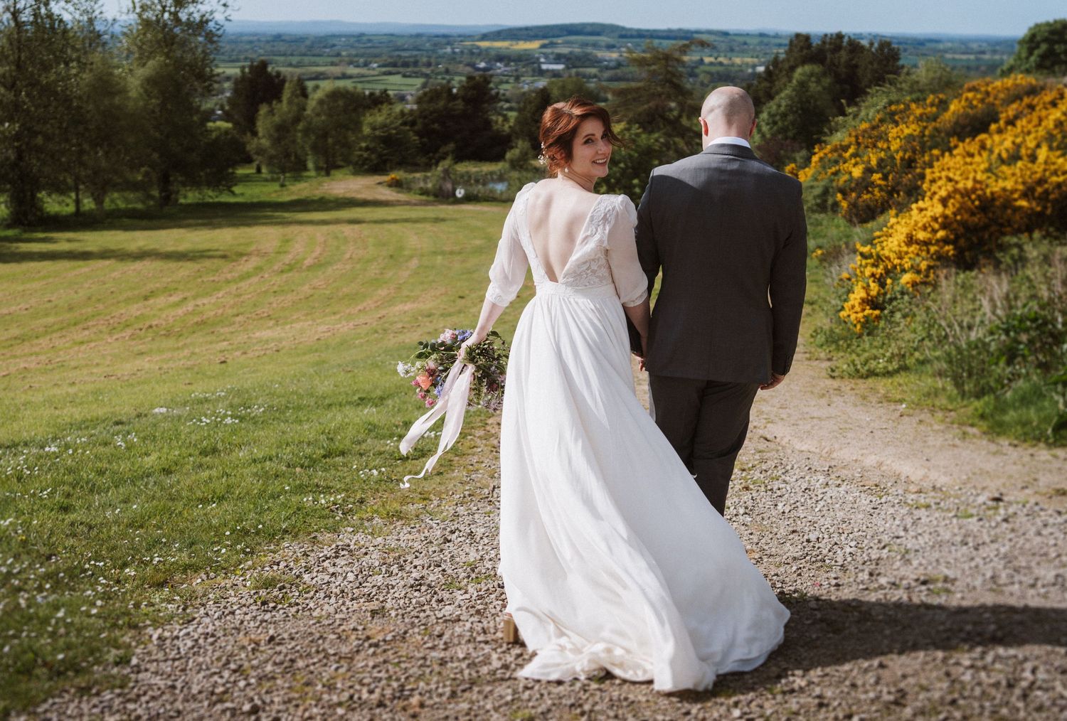 Bride in white gown holding bouquet walks with groom on gravel path through scenic Irish countryside.