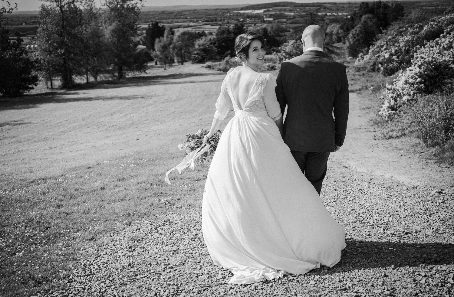 Black and white photo of a bride and groom walking hand in hand on a gravel path surrounded by lush greenery.