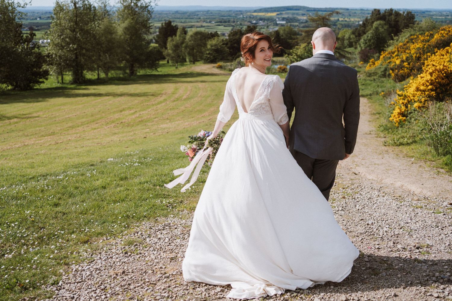 A bride in a white gown and groom in a grey suit walk hand in hand down a gravel path surrounded by lush green countryside.