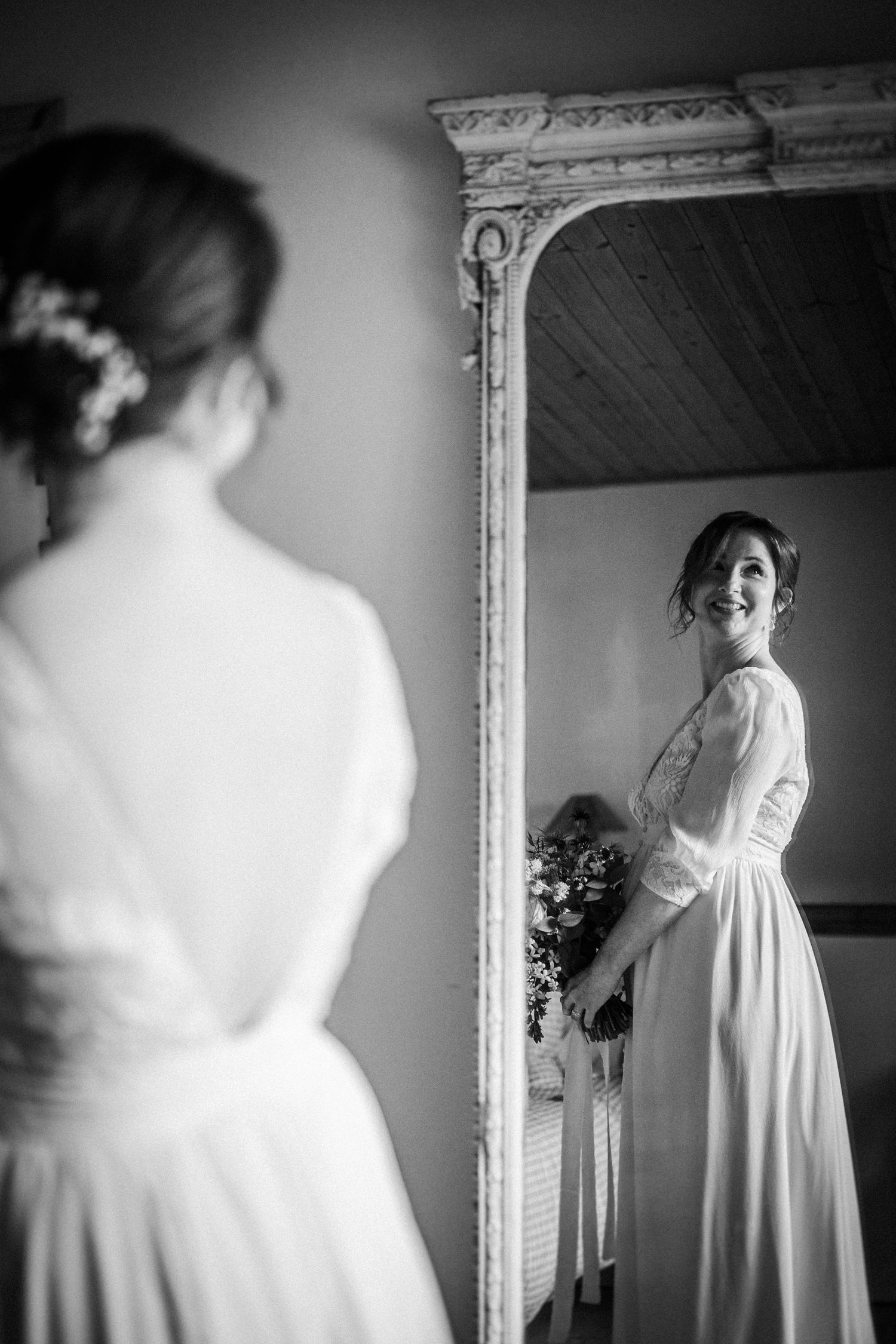 Bride in white gown smiling at her reflection in an ornate mirror, captured in elegant black and white photography.