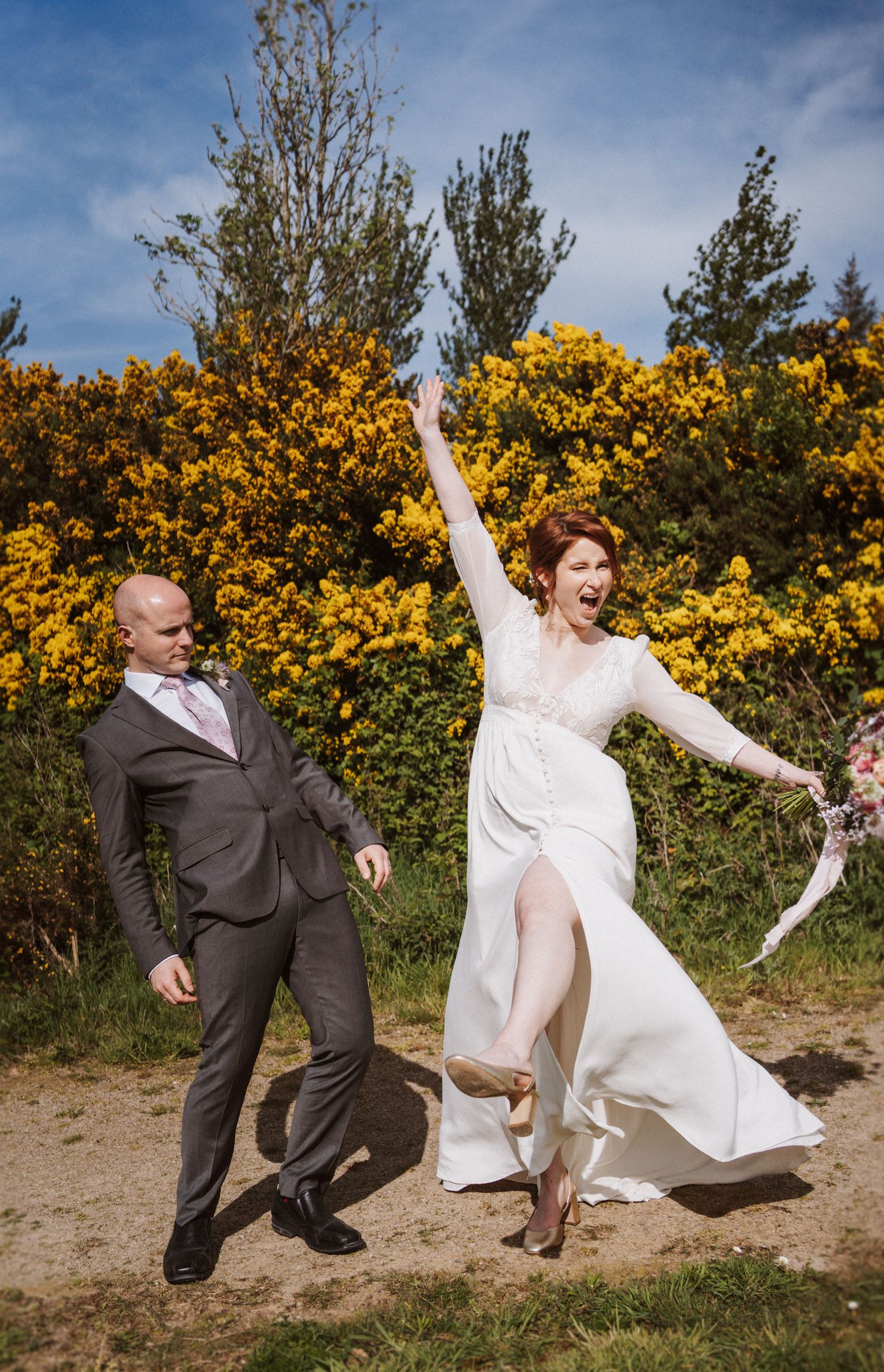 Joyful bride in white dress raises arm celebrating with groom in gray suit, yellow gorse bushes backdrop.