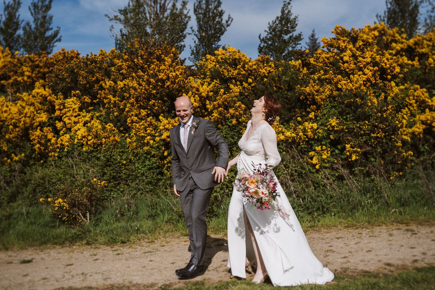 Bride and groom walking outdoors near yellow flowering bushes, bride holding colorful bouquet in white gown.