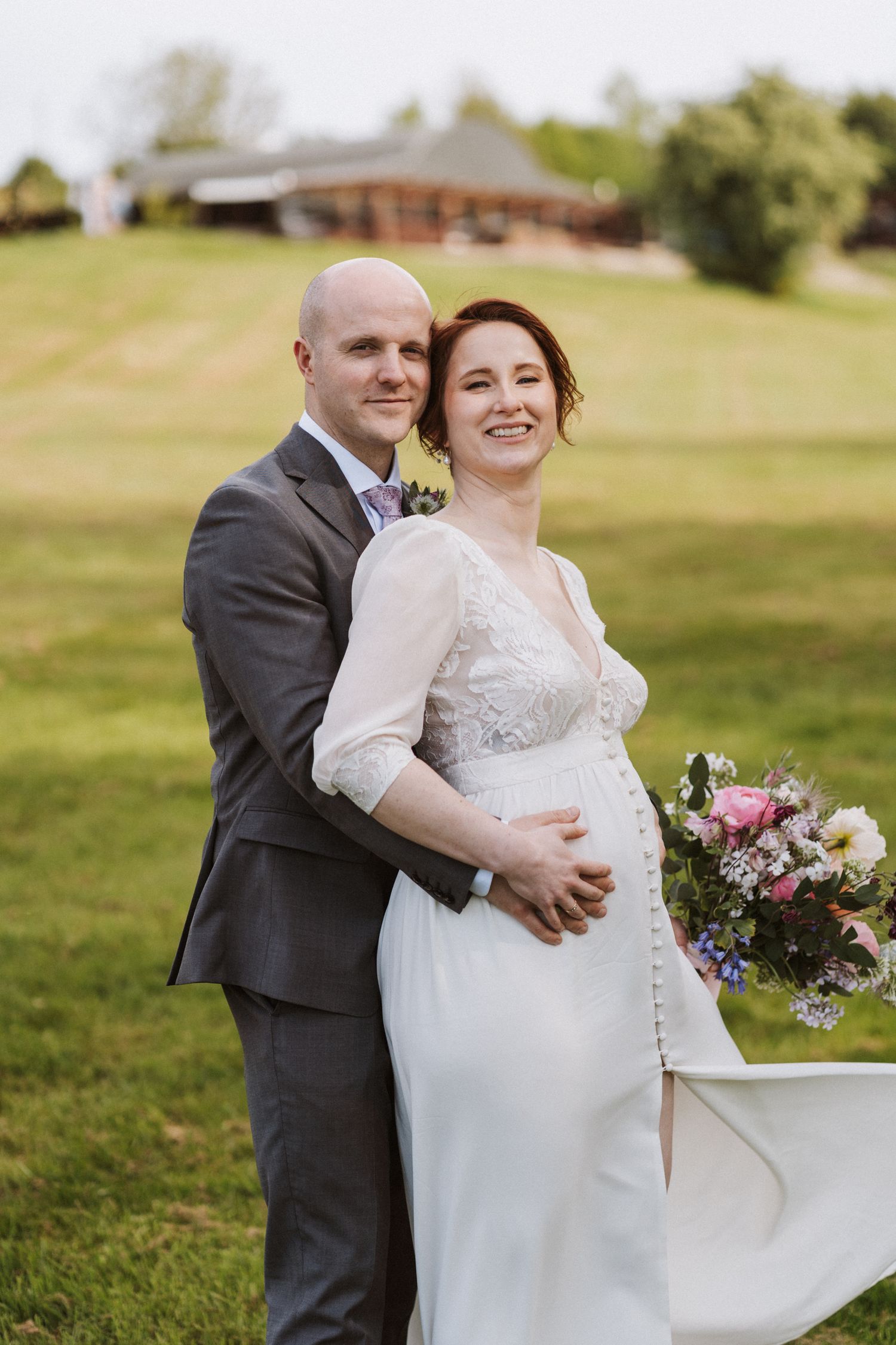 Bride and groom pose together outdoors on green lawn, bride holding colorful bouquet in elegant white wedding gown.