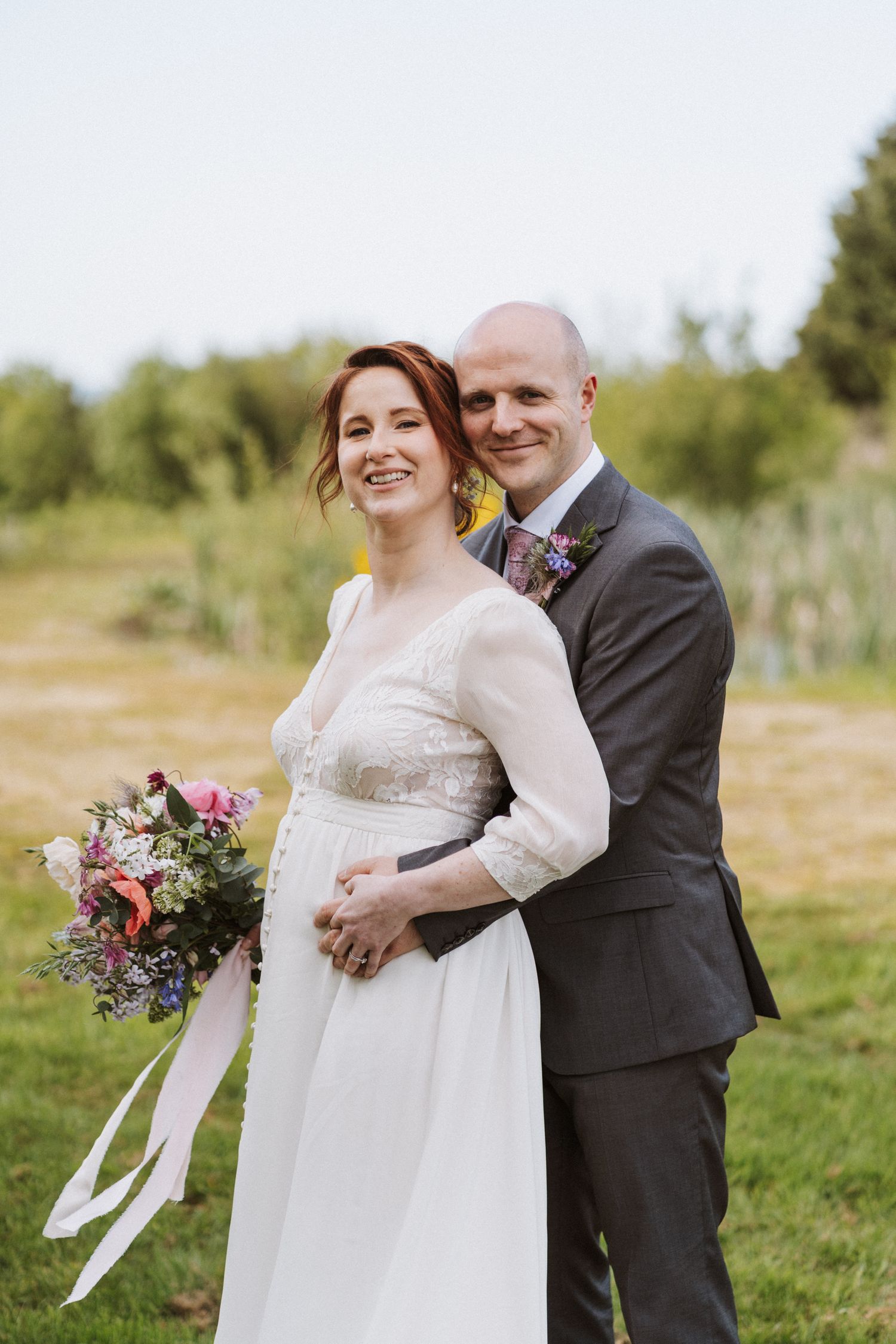 Smiling bride holding colorful bouquet with ribbons stands with groom in gray suit outdoors at wedding.