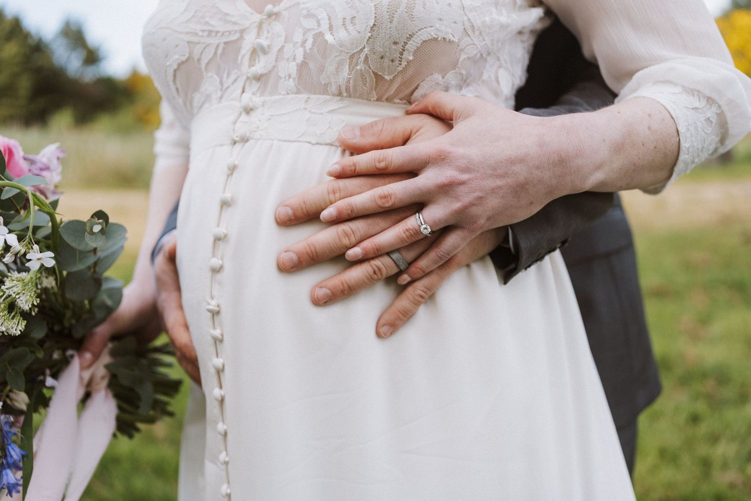 Pregnant bride in white lace gown with groom's hands on baby bump, wearing wedding rings, holding floral bouquet outdoors.
