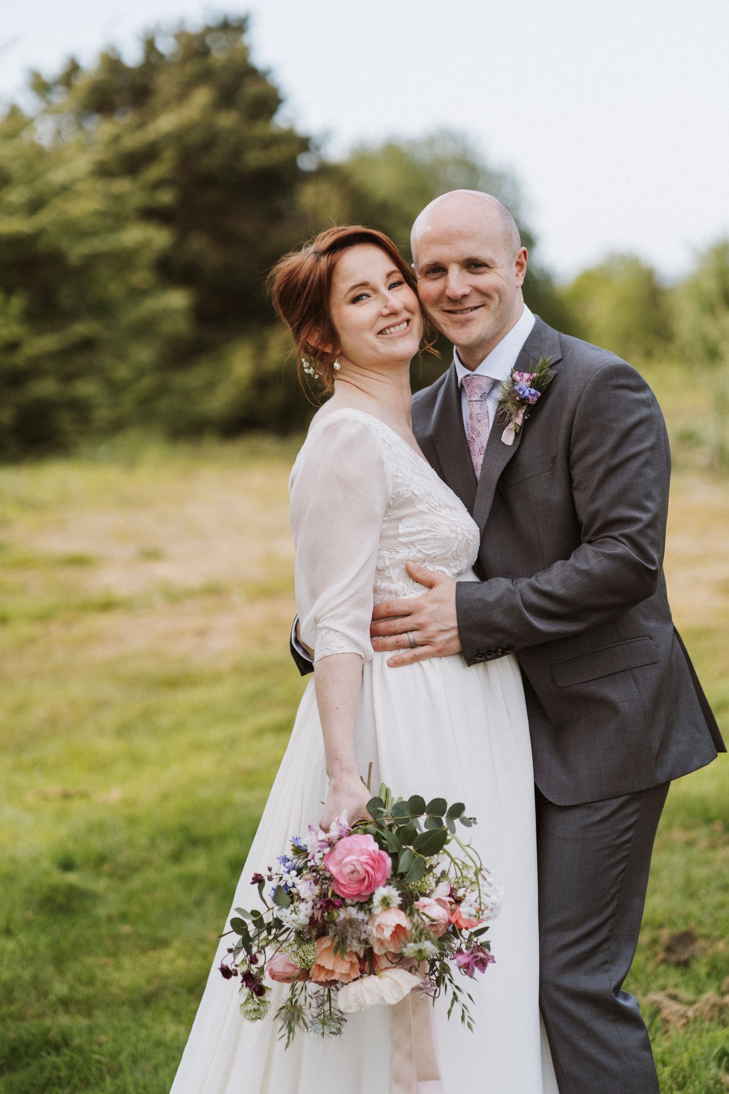 Bride and groom embrace outdoors, she holds a pink floral bouquet, wearing white gown, he wears a gray suit.