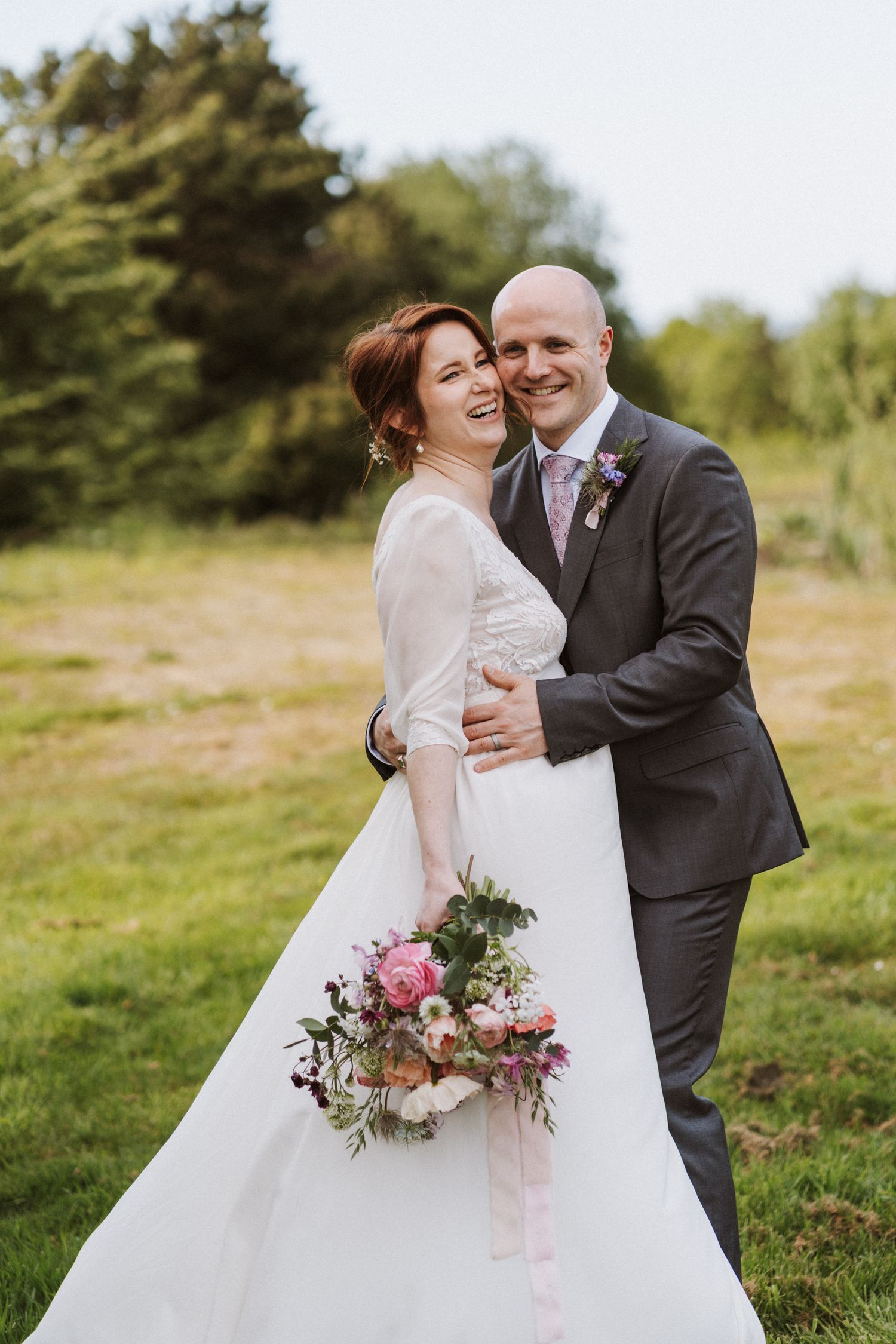 Happy bride and groom embracing outdoors, bride holding pink floral bouquet, groom in gray suit, green field background.