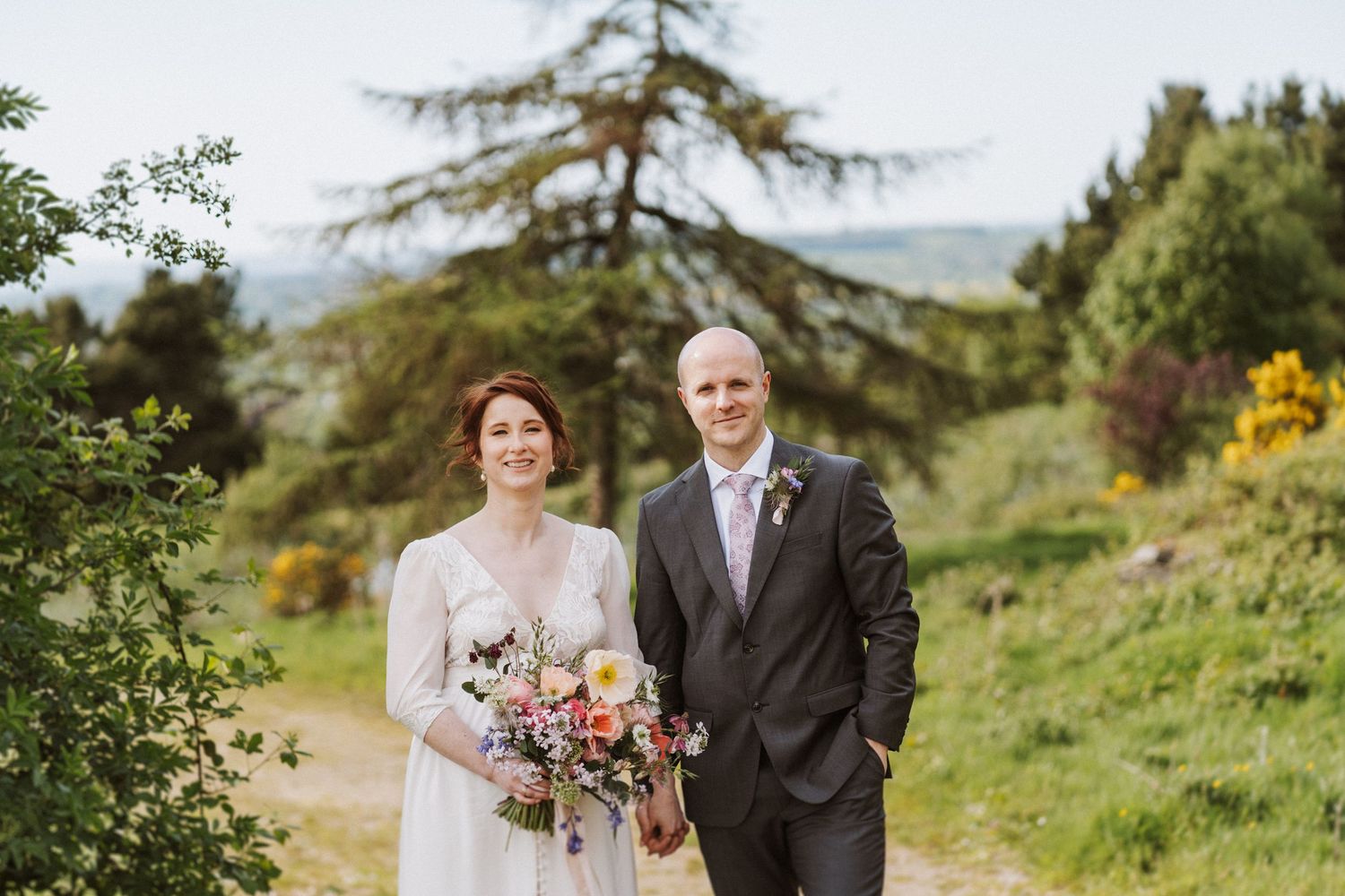 Bride and groom pose outdoors on a garden path, bride holding colorful wildflower bouquet, surrounded by lush greenery.