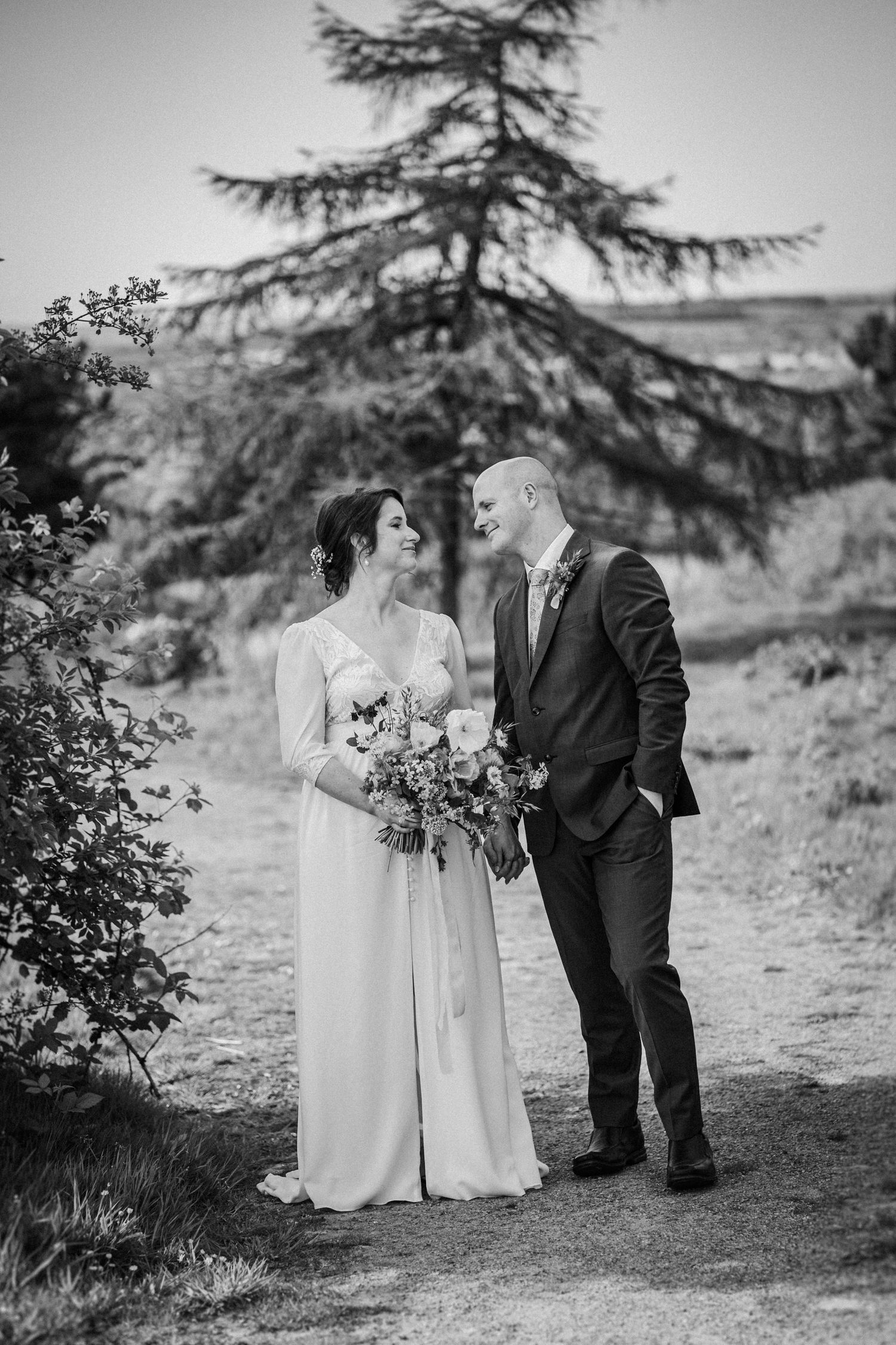 Black and white photo of a bride and groom sharing a loving glance on a garden path, bride holding a floral bouquet.