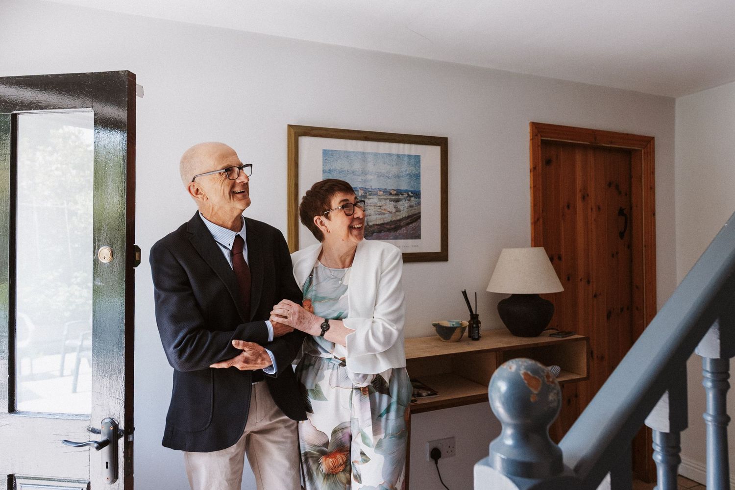 Elderly couple in formal attire standing together indoors near a staircase, smiling and conversing in a bright home.