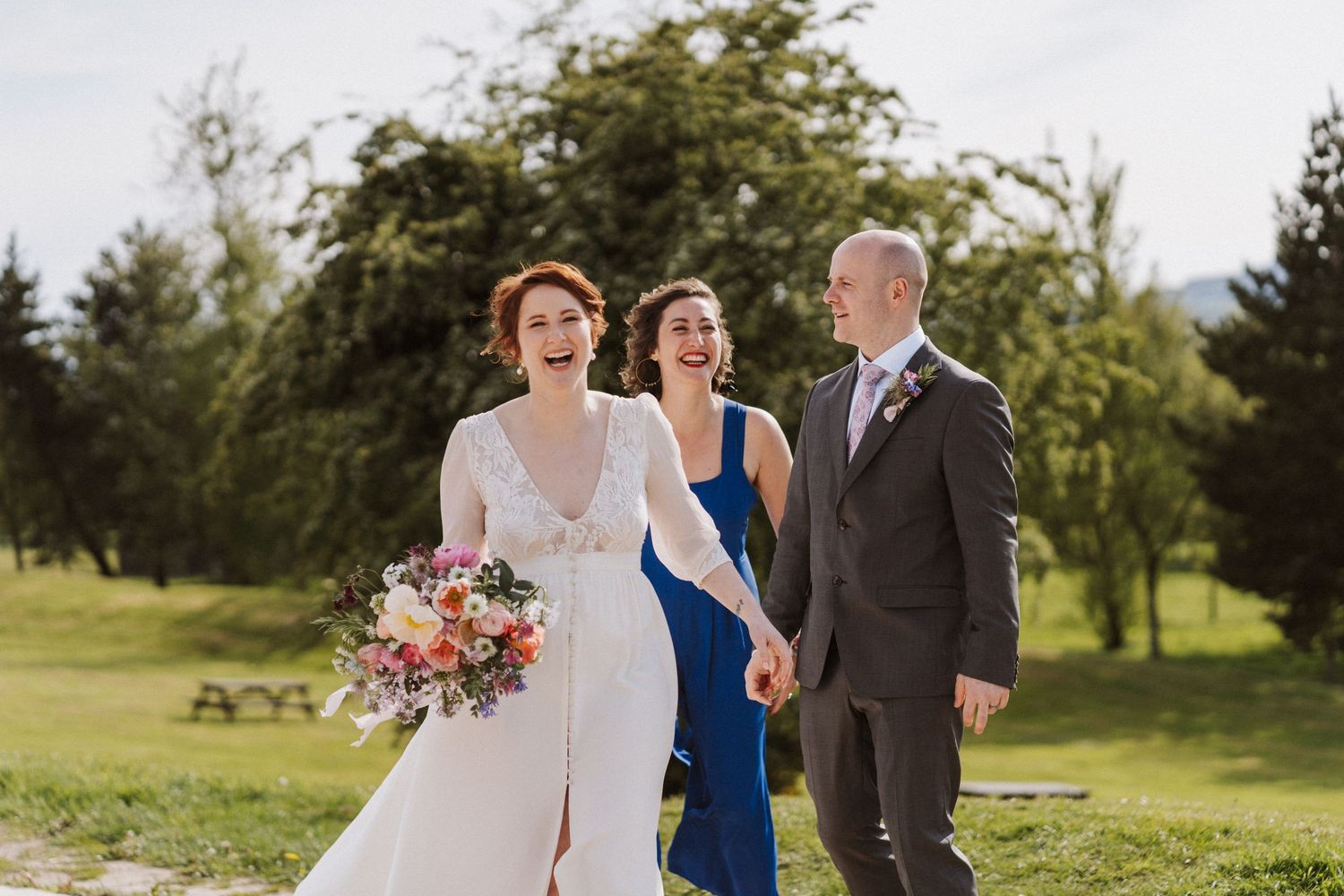Bride in white gown laughing with bridesmaid in blue dress and groom in suit walking outdoors at wedding.