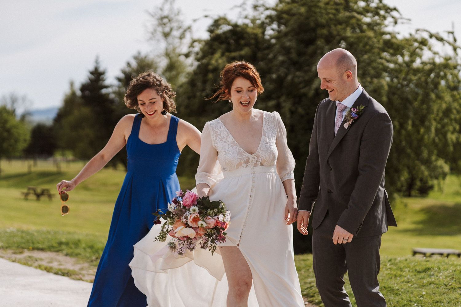 Bride in white gown walks with bridesmaid in blue dress and groomsman in gray suit on sunny outdoor wedding path.
