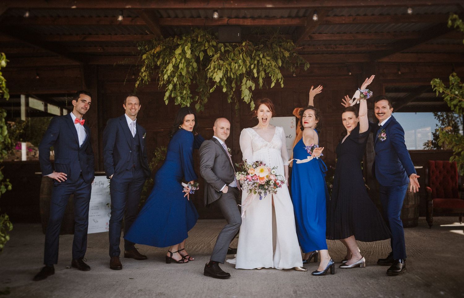 Wedding party posing joyfully outdoors under hanging greenery, bride in white gown holding colorful bouquet.