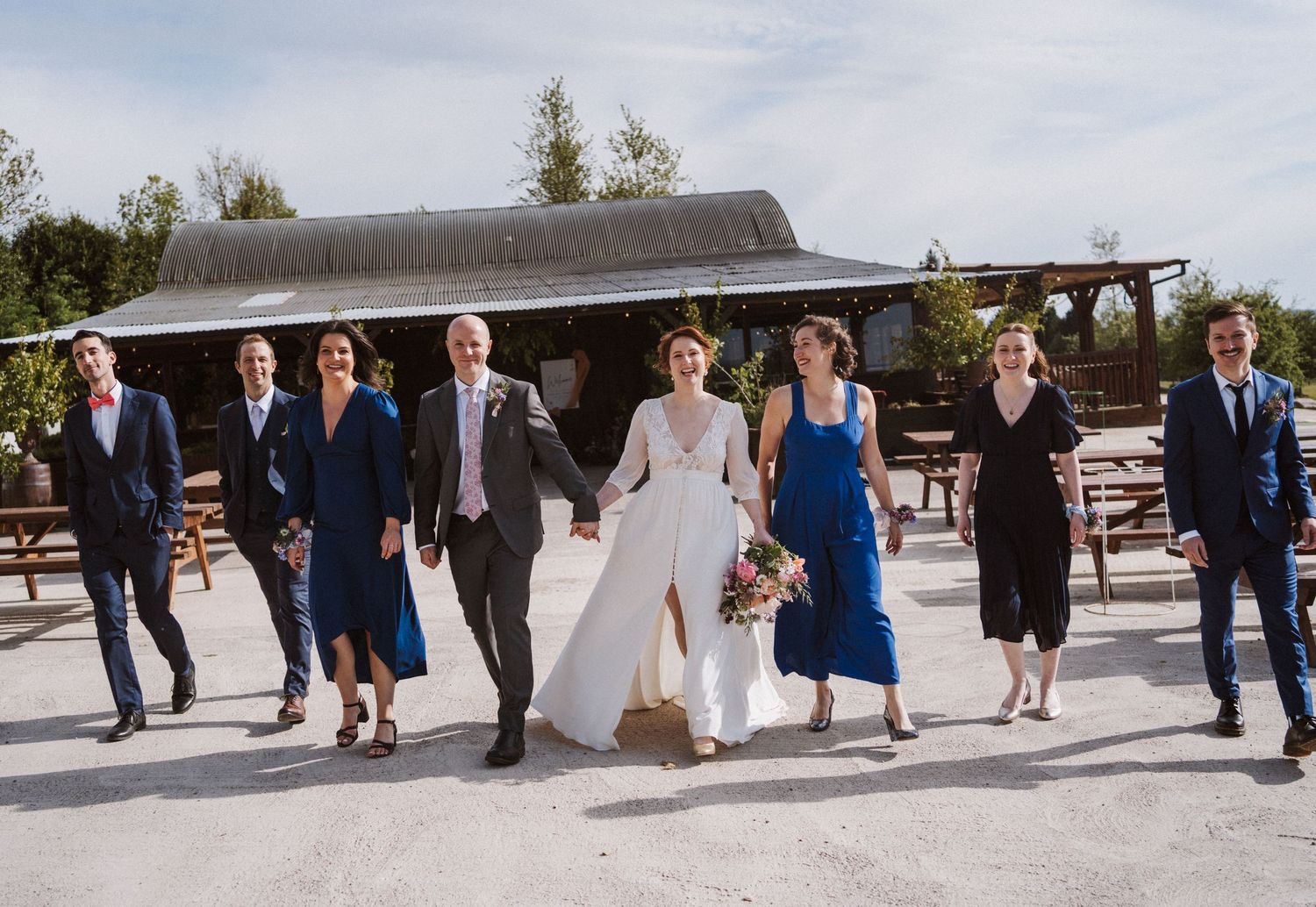 Wedding party walking together outdoors, bride in white gown with groomsmen in navy suits and bridesmaids in blue dresses.