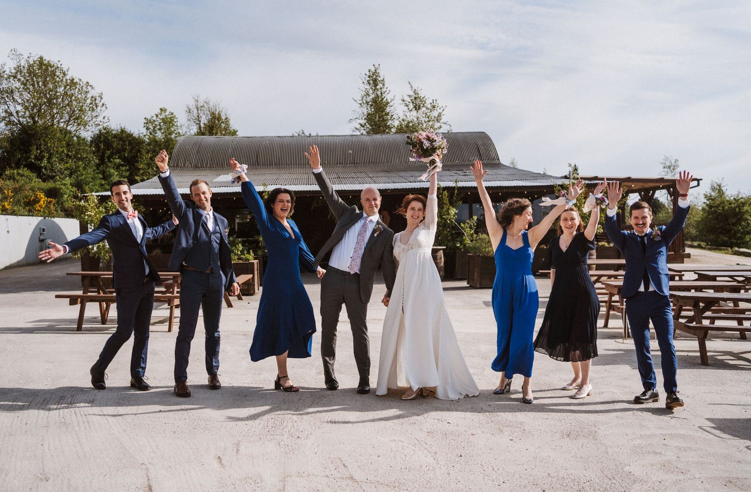 Wedding party celebrating outdoors with arms raised, bride in white gown, guests in navy and blue attire, rustic venue backdrop.