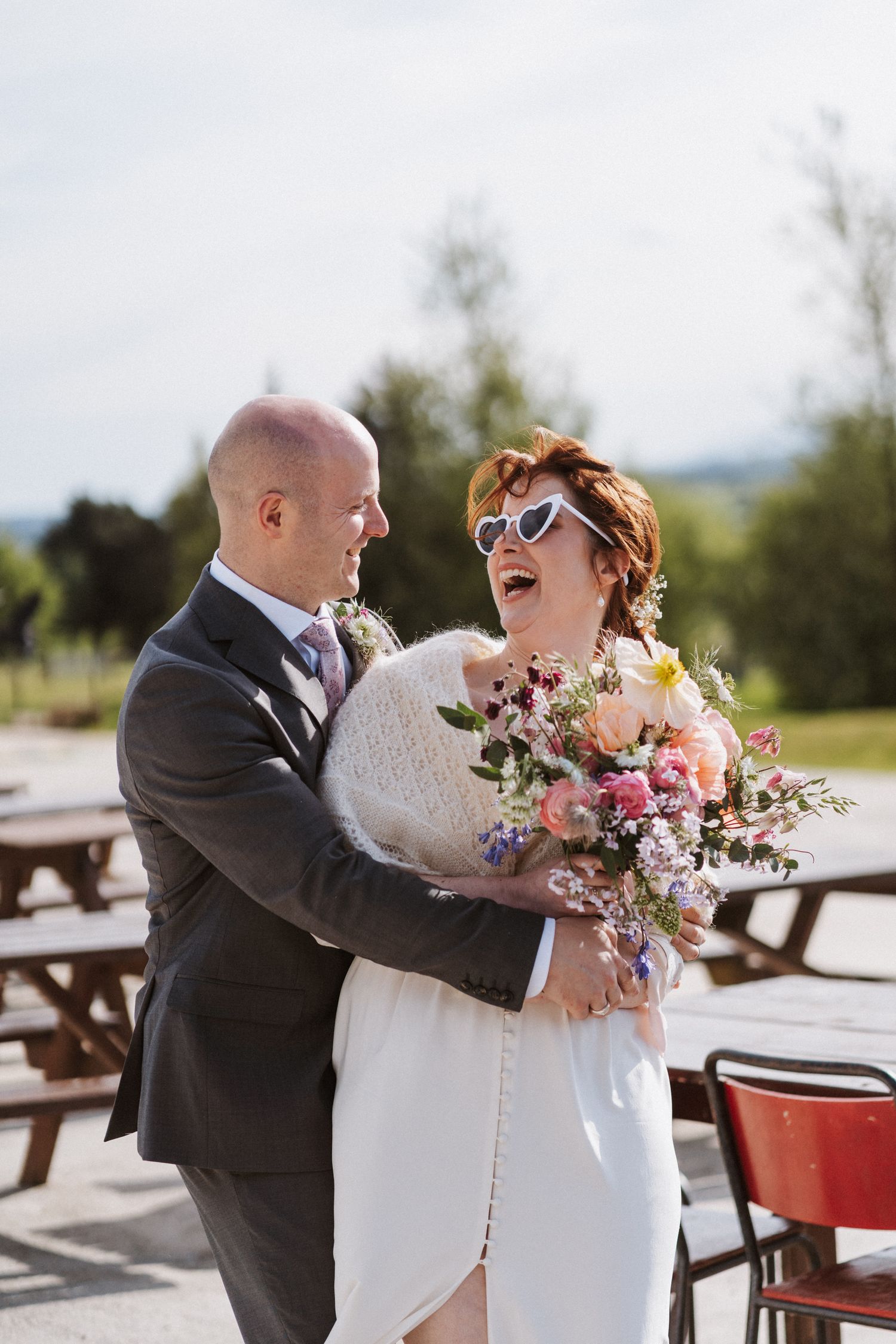 A laughing bride in heart sunglasses holds a colorful bouquet while embracing a groom in a gray suit outdoors.