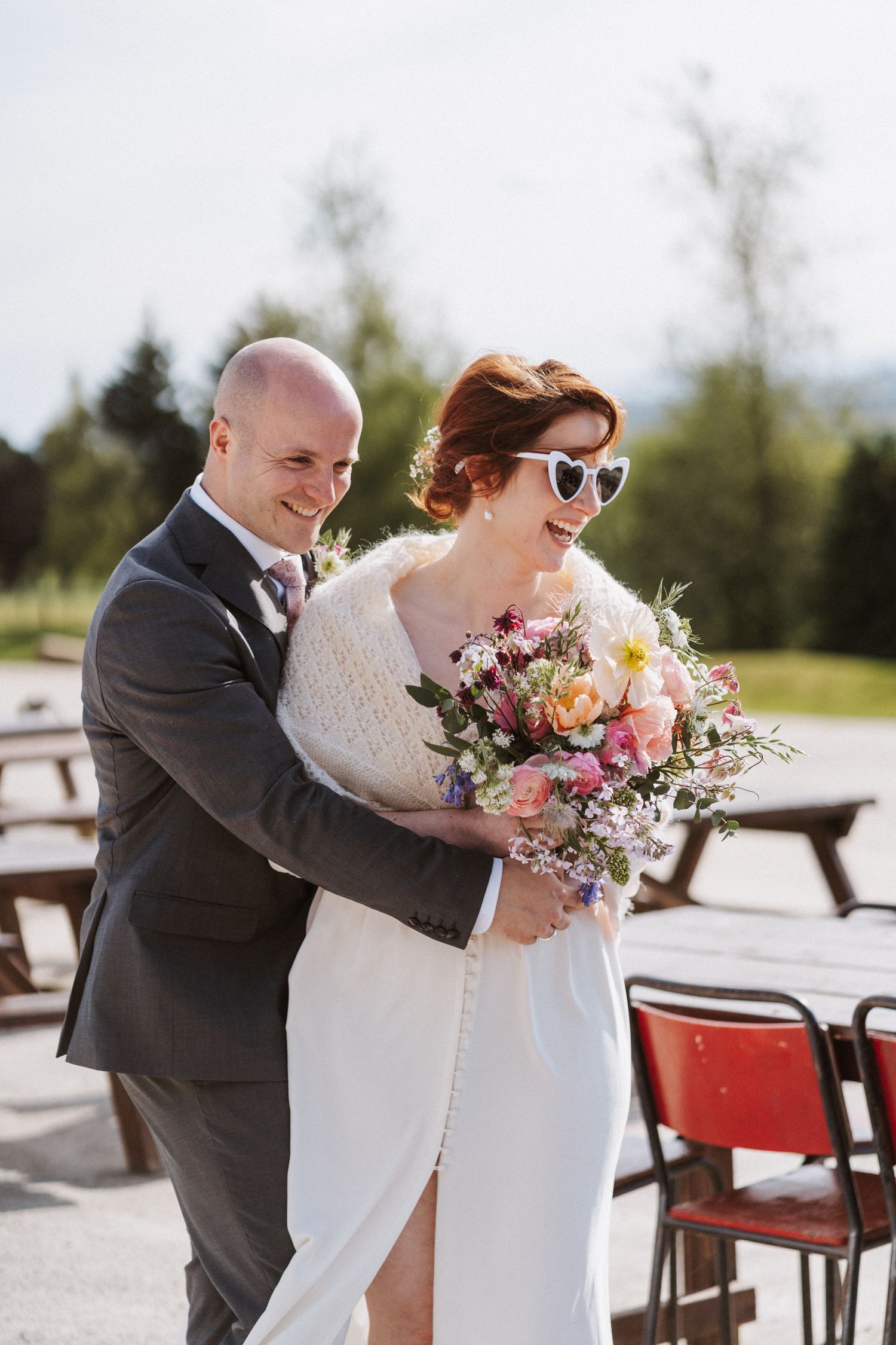 Bride in white dress and heart sunglasses holds colorful bouquet while groom in gray suit embraces her outdoors.