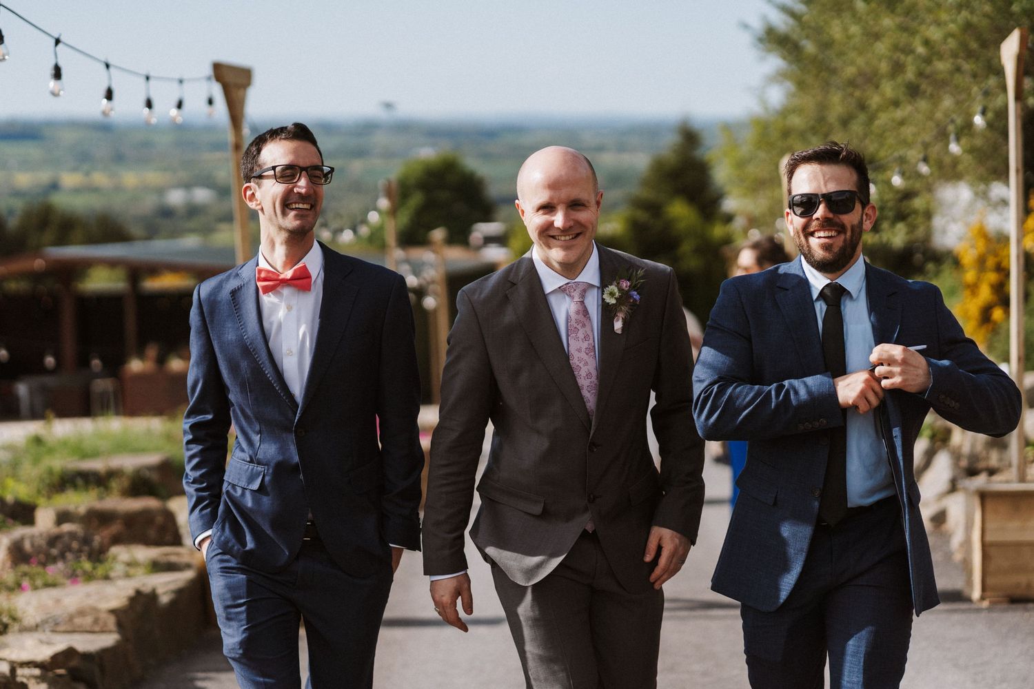 Three well-dressed groomsmen in navy suits walking together outdoors at a wedding venue on a sunny day.