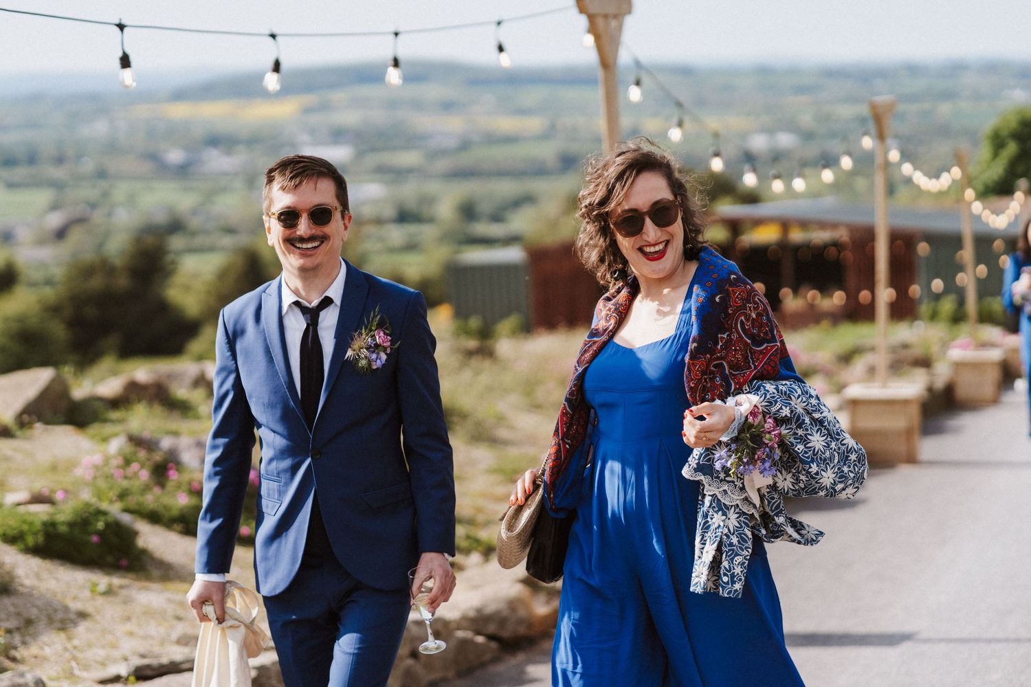 A smiling couple in blue attire walks outdoors at a wedding venue with scenic countryside views and festoon lights.