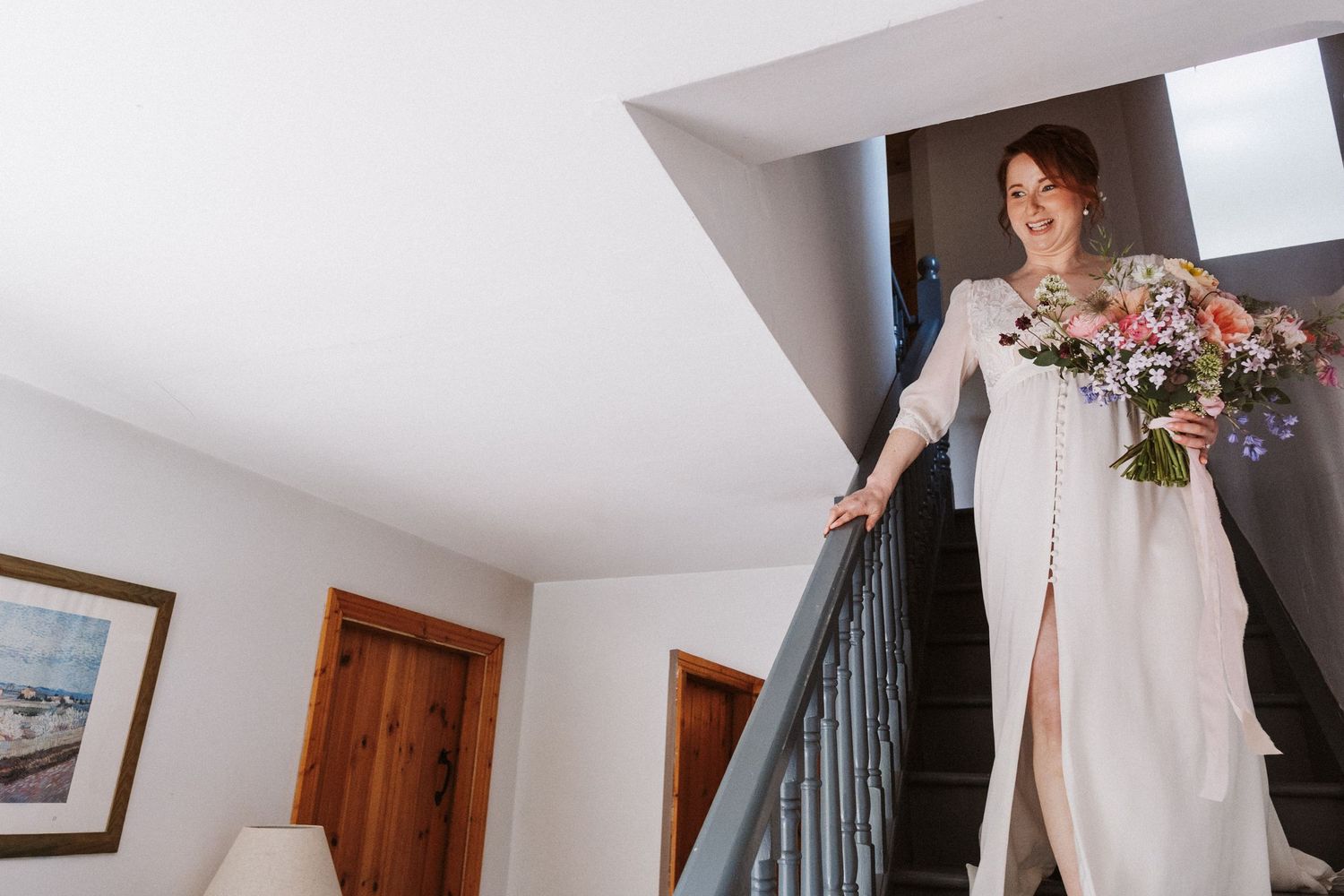 Bride in white gown holding colorful bouquet descends staircase, smiling, on her wedding day.